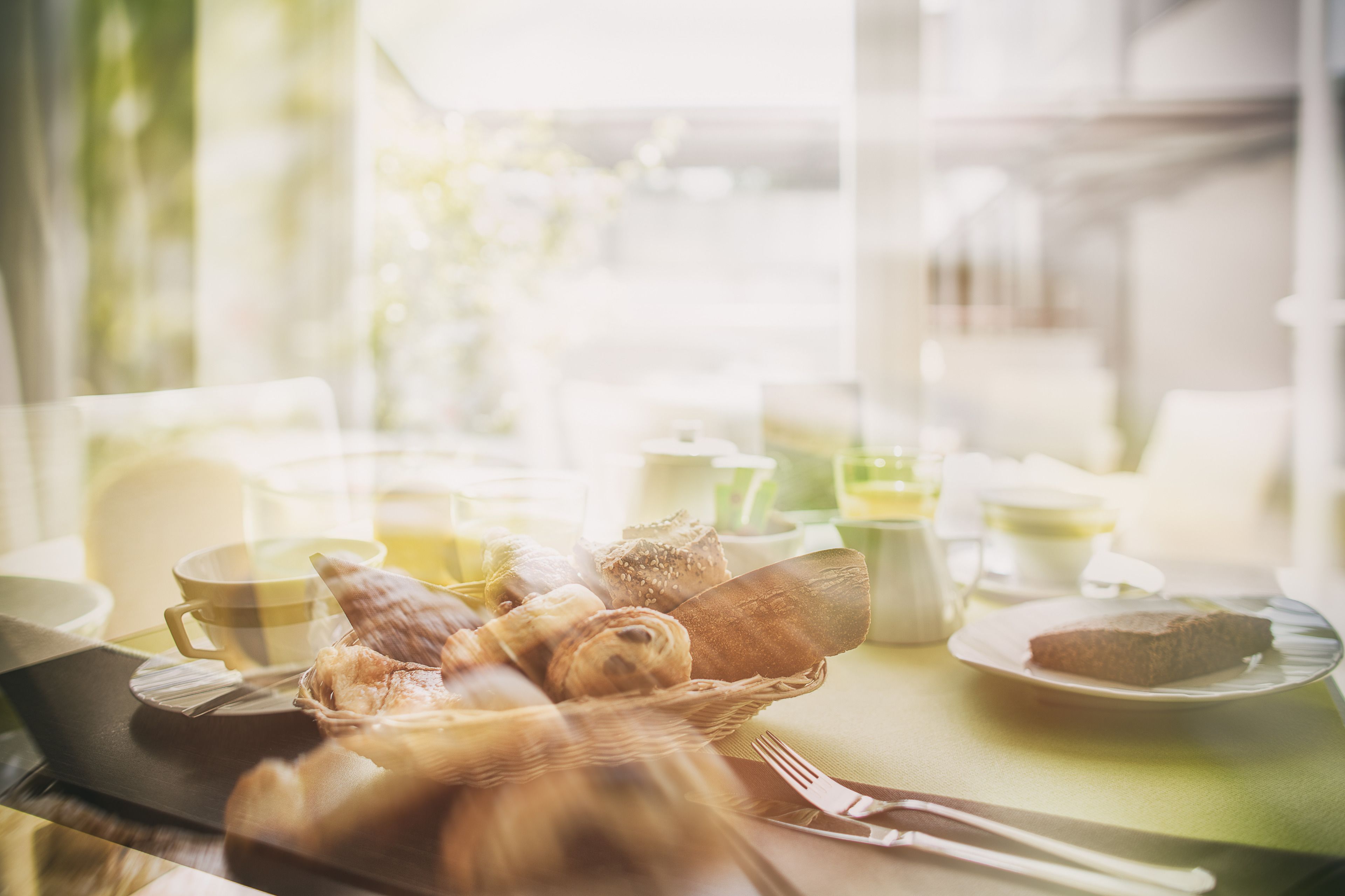 A breakfast setting with bread, pastries, and a slice of cake on a plate, with cups and a teapot in the background.