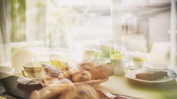 A breakfast setting with bread, pastries, and a slice of cake on a plate, with cups and a teapot in the background.