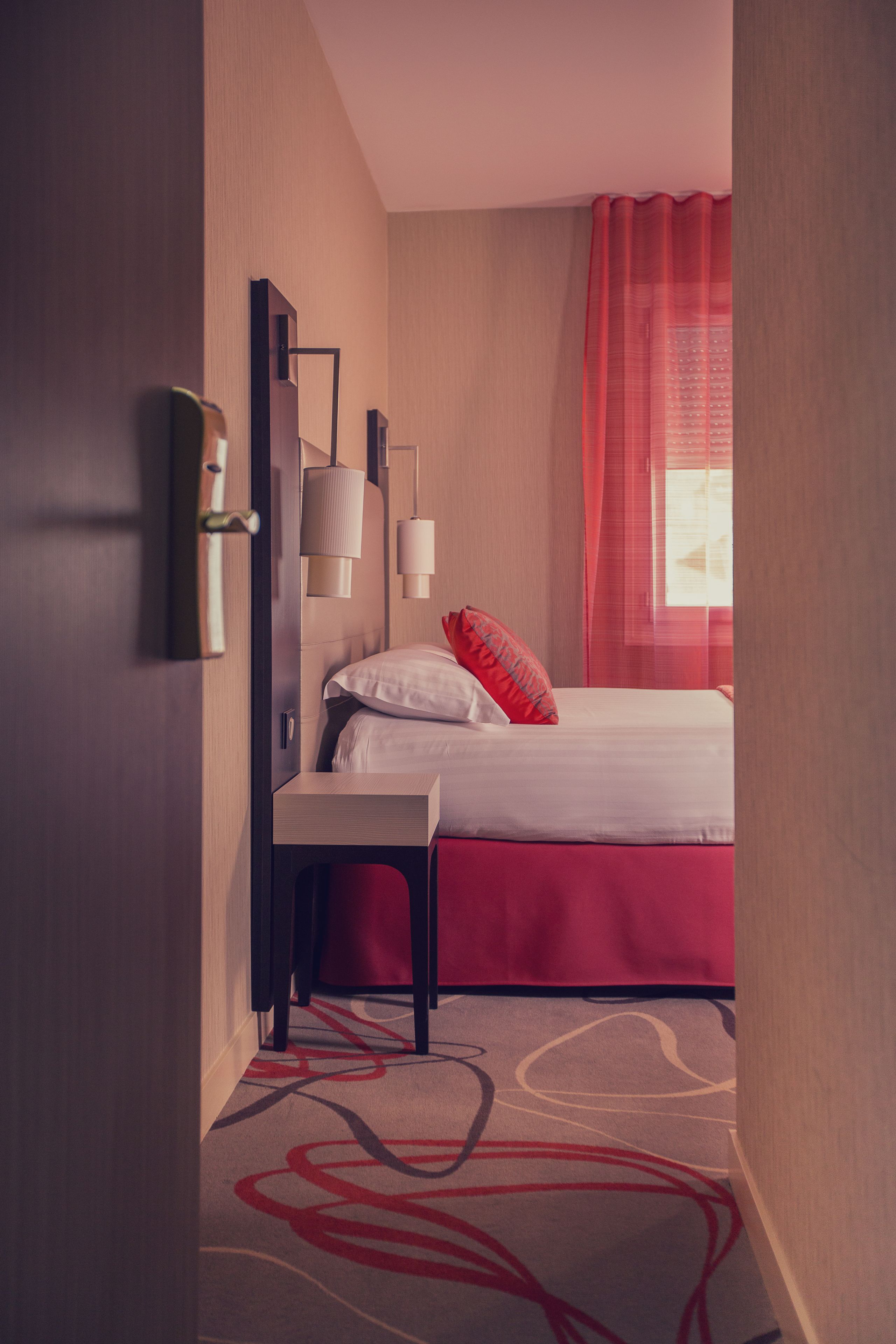A hotel room with a bed, a bedside table, a lamp, and a window with curtains.