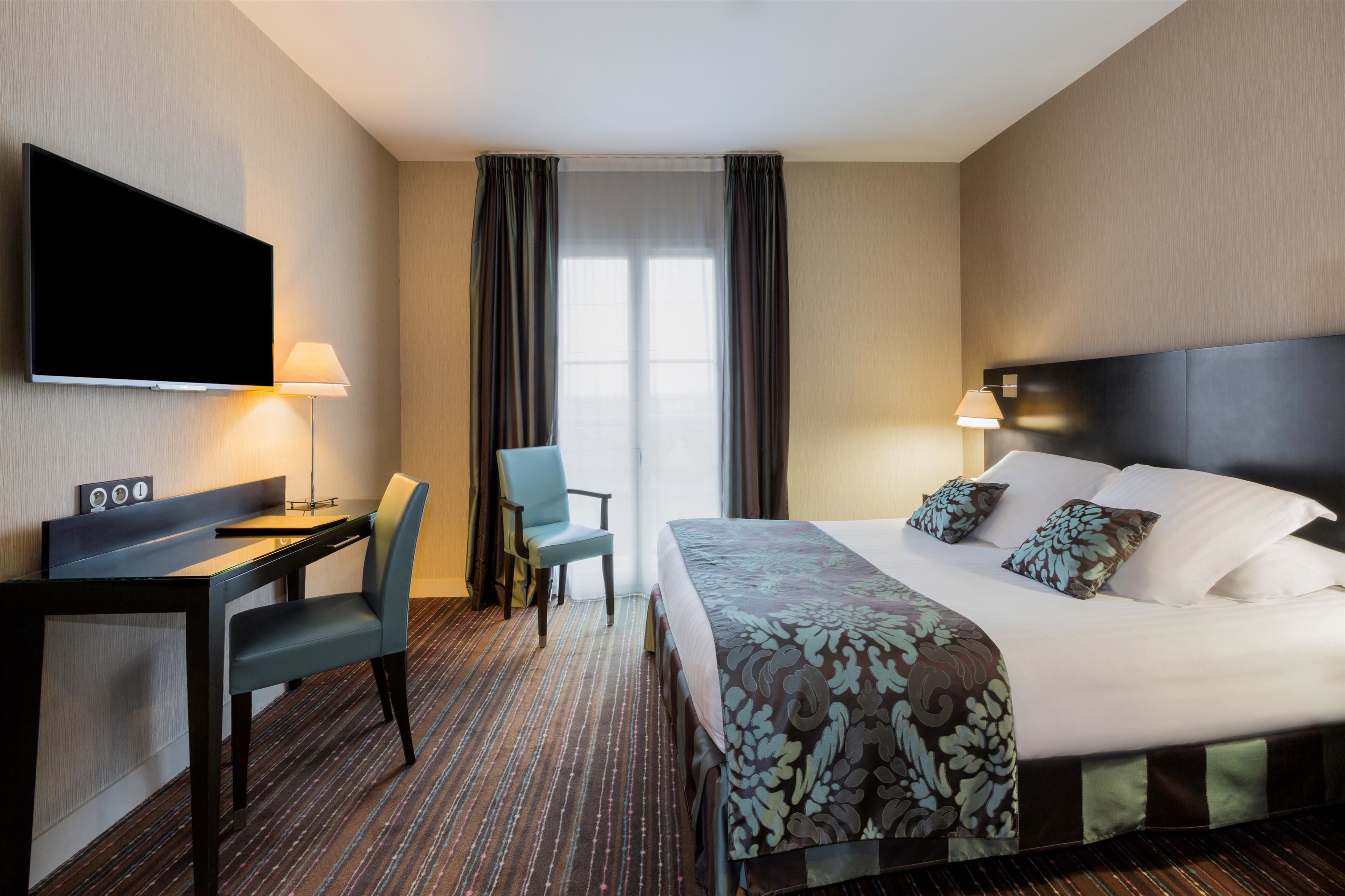 A hotel room with a bed, a desk, a chair, a TV, and a window with curtains.