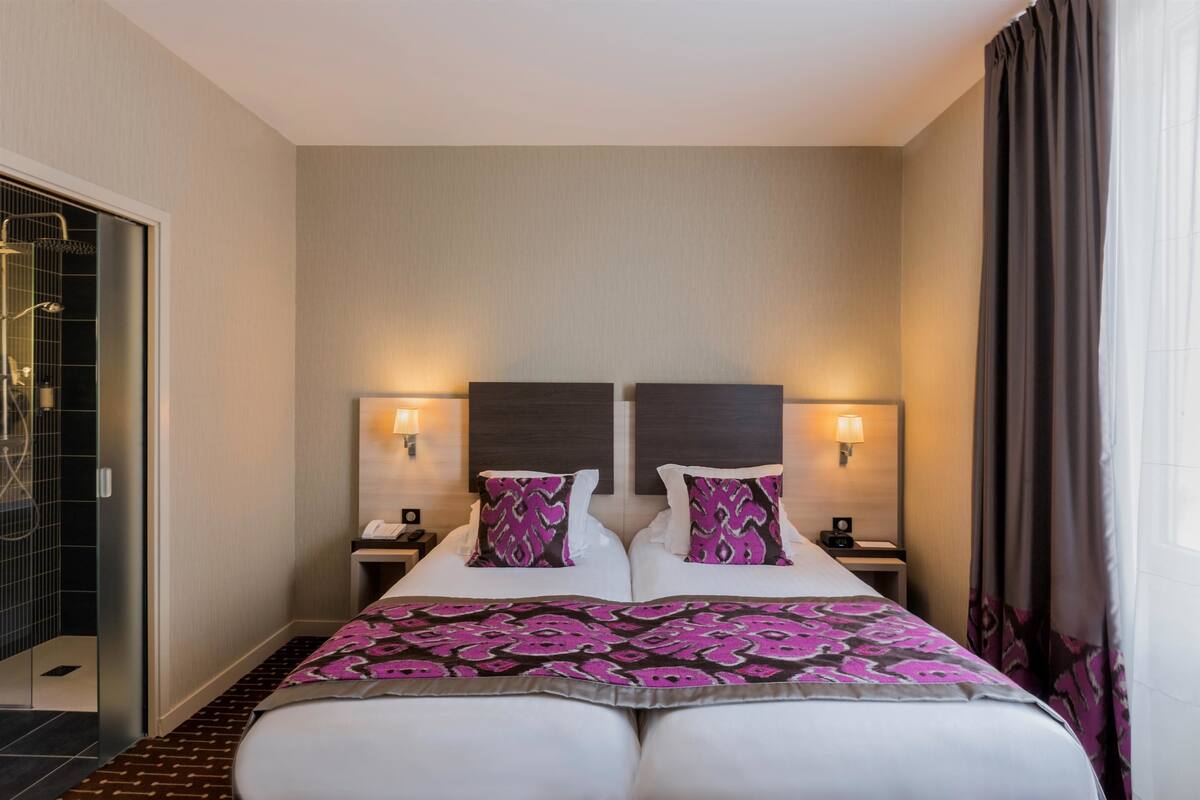A double bed with a patterned bedspread, two bedside tables with lamps, and a shower area visible to the left.