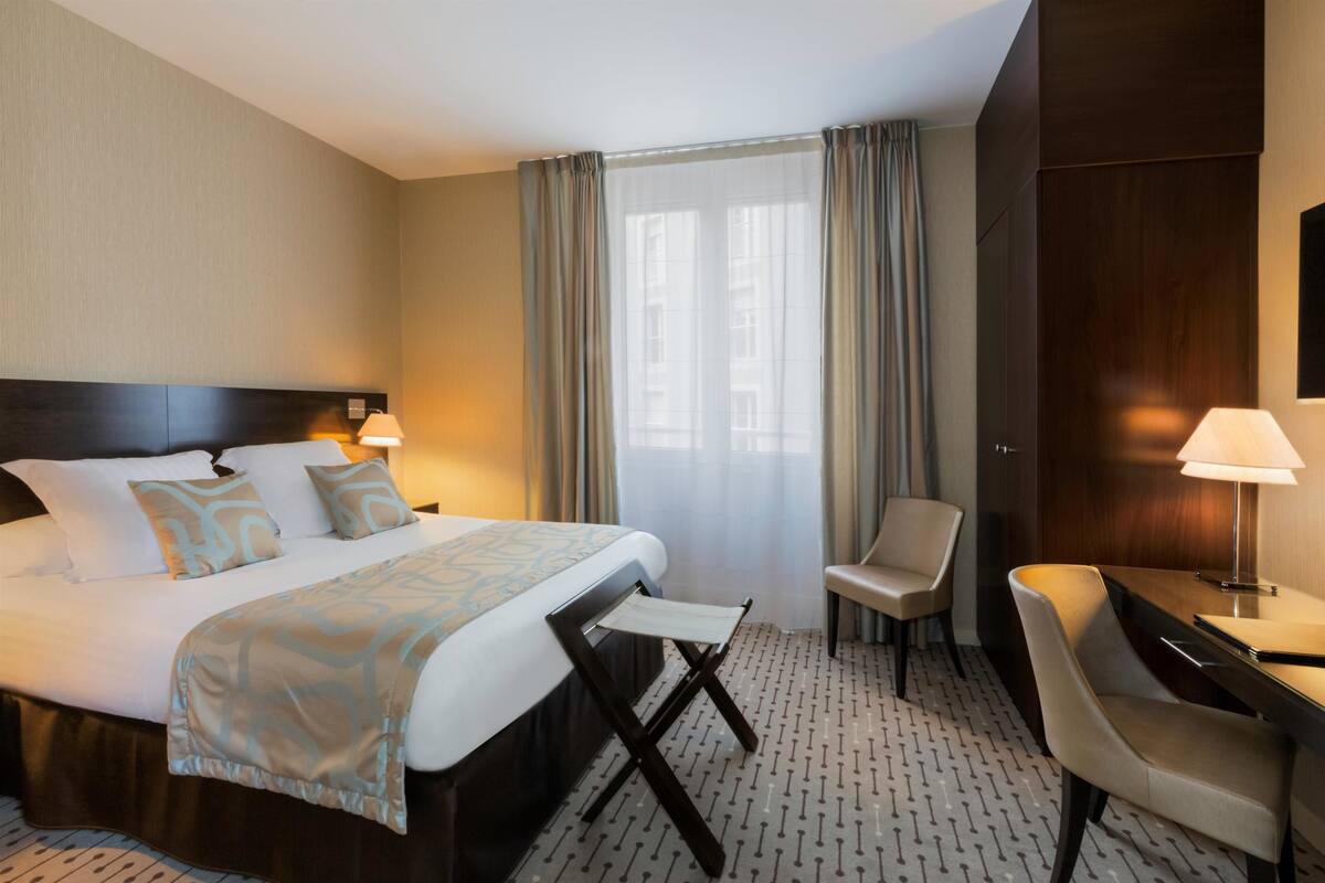 A hotel room with a large bed, a desk, a chair, and a window with curtains.