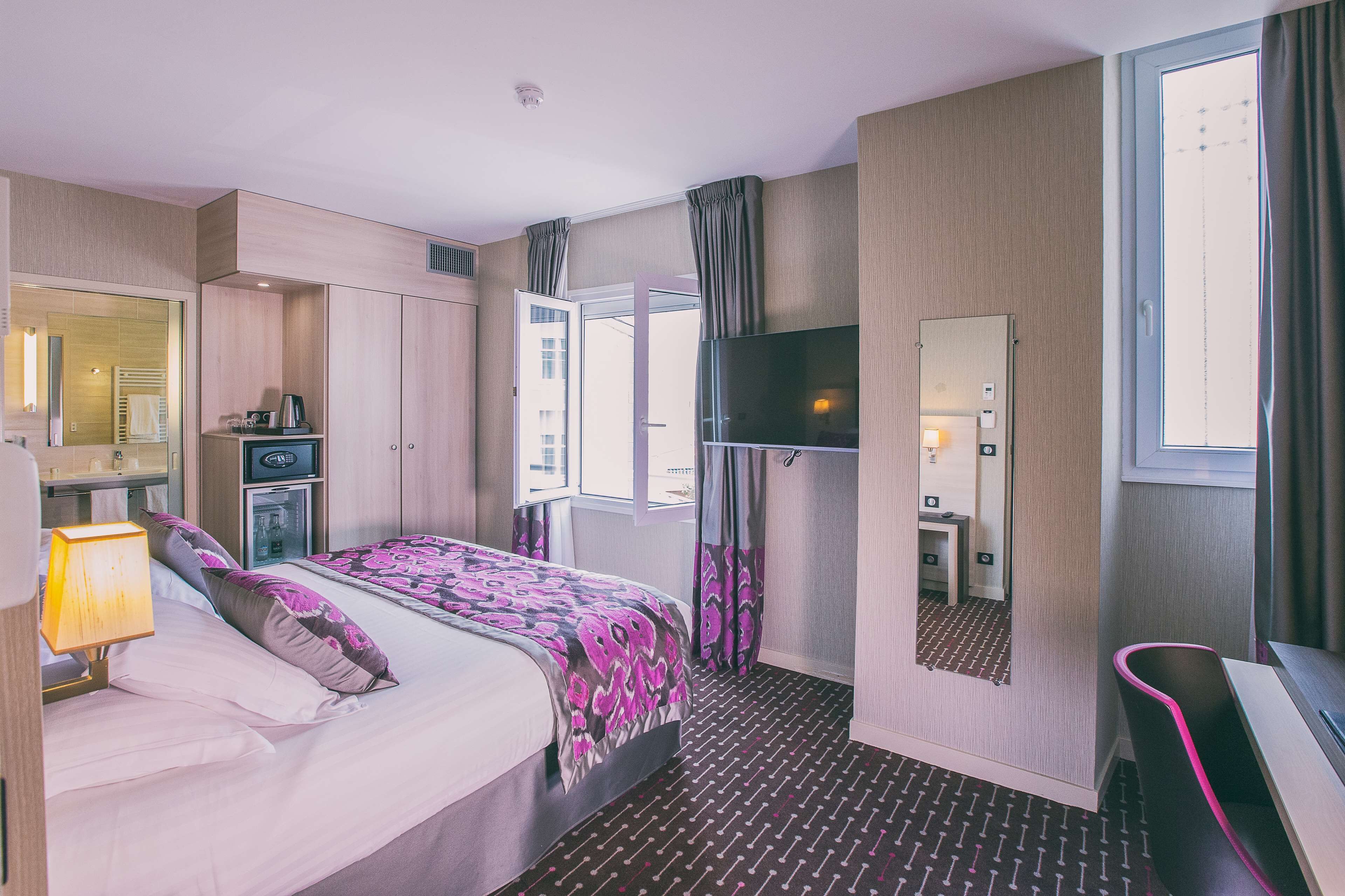 A hotel room with a bed, a desk, a TV, a mirror, and a window with curtains.