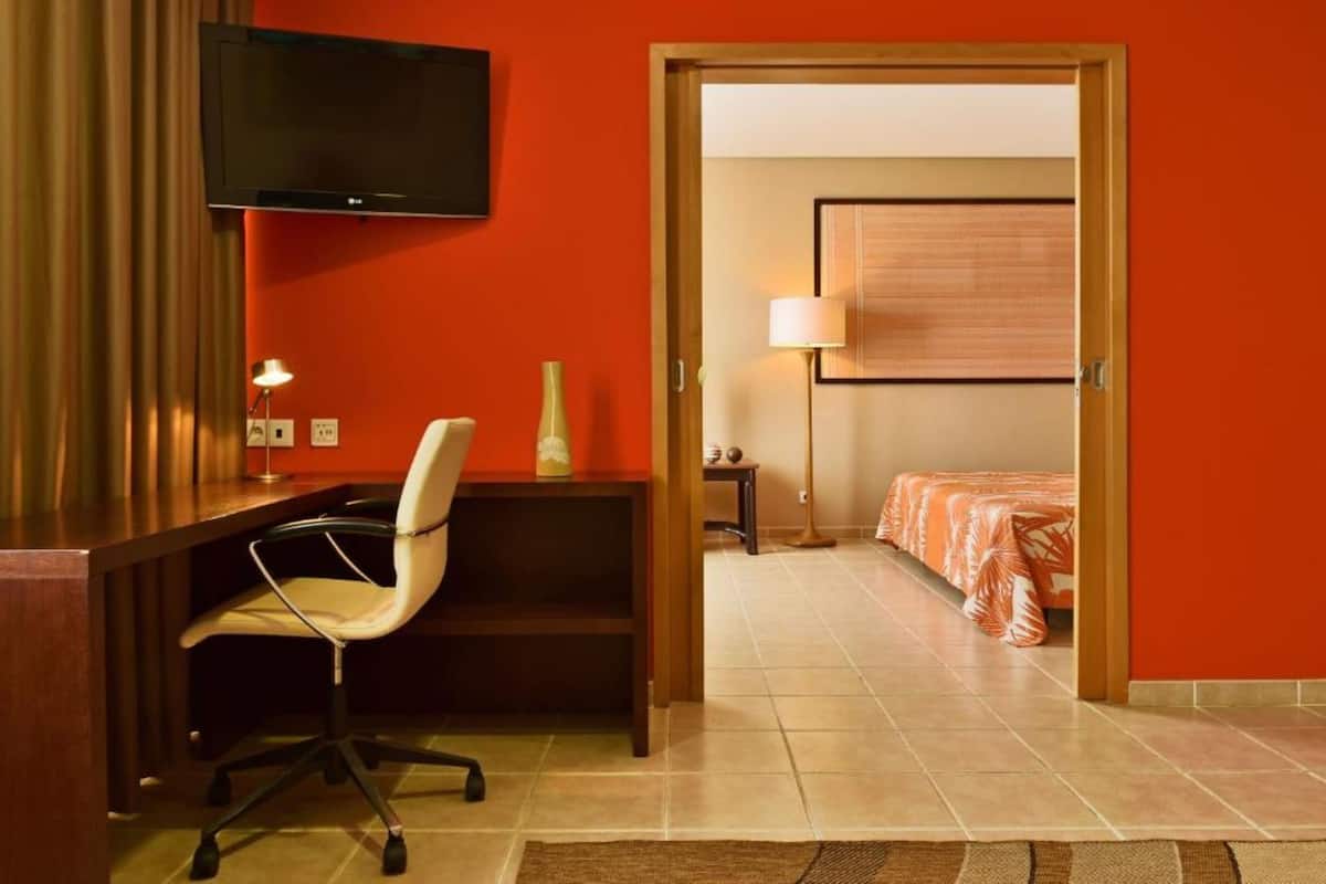Een hotelkamer met een bureau, stoel, televisie, bed en een deur naar een andere kamer.