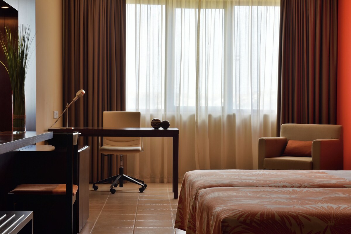 Une chambre d’hôtel avec un lit, un bureau et une chaise, une lampe et une plante en pot.