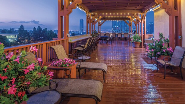 Un espacio de estar al aire libre cubierto con muebles de madera y luces colgantes, con vistas a un paisaje urbano.