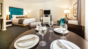 Una habitación de hotel con una cama bien hecha, una mesa de comedor dispuesta para dos personas con copas de vino y cubiertos, un televisor y un minibar.