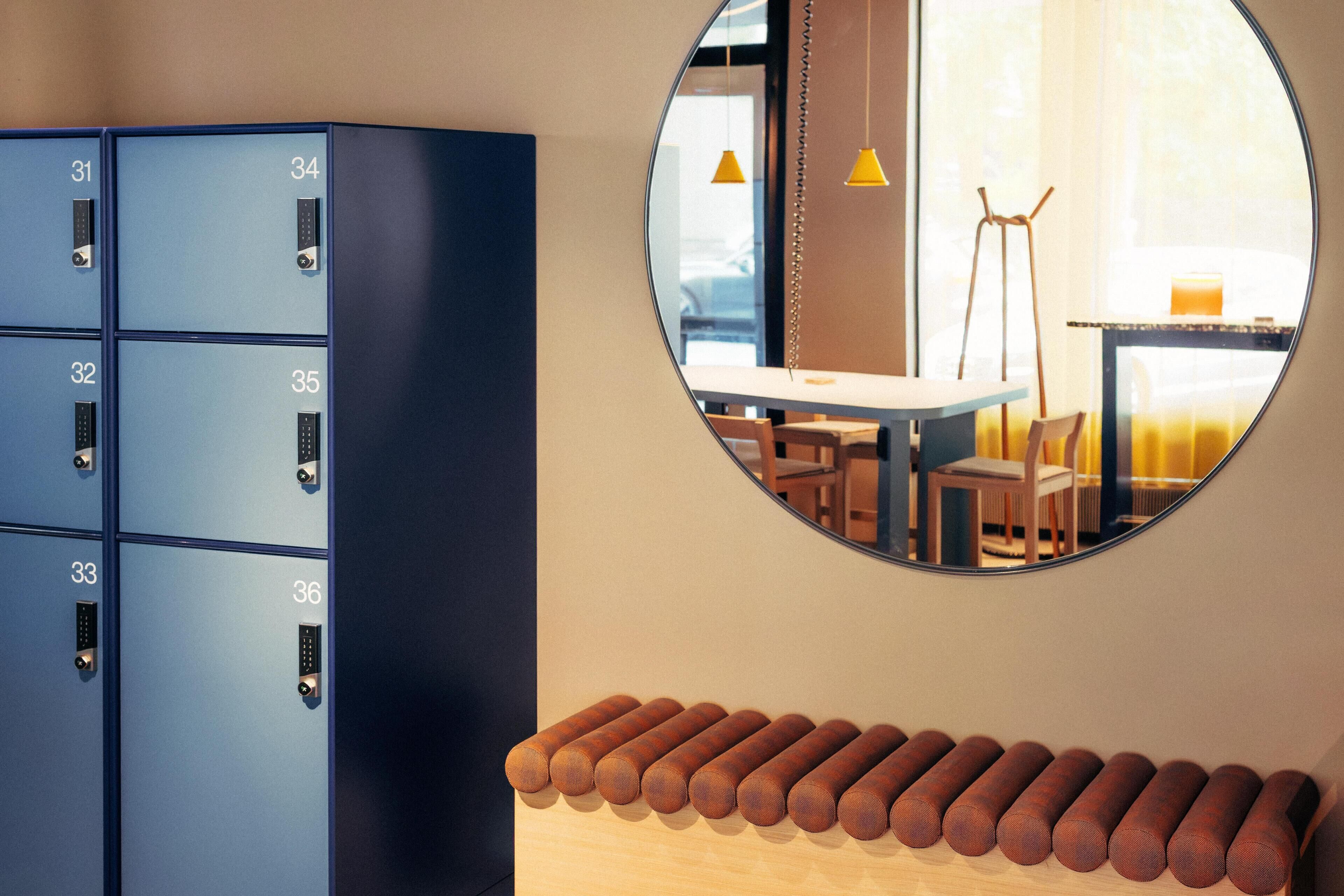 Een blauwe opslagunit met genummerde vakken, een ronde spiegel die een kamer met een tafel en stoelen weerspiegelt, en een houten bank met cilindervormige objecten.