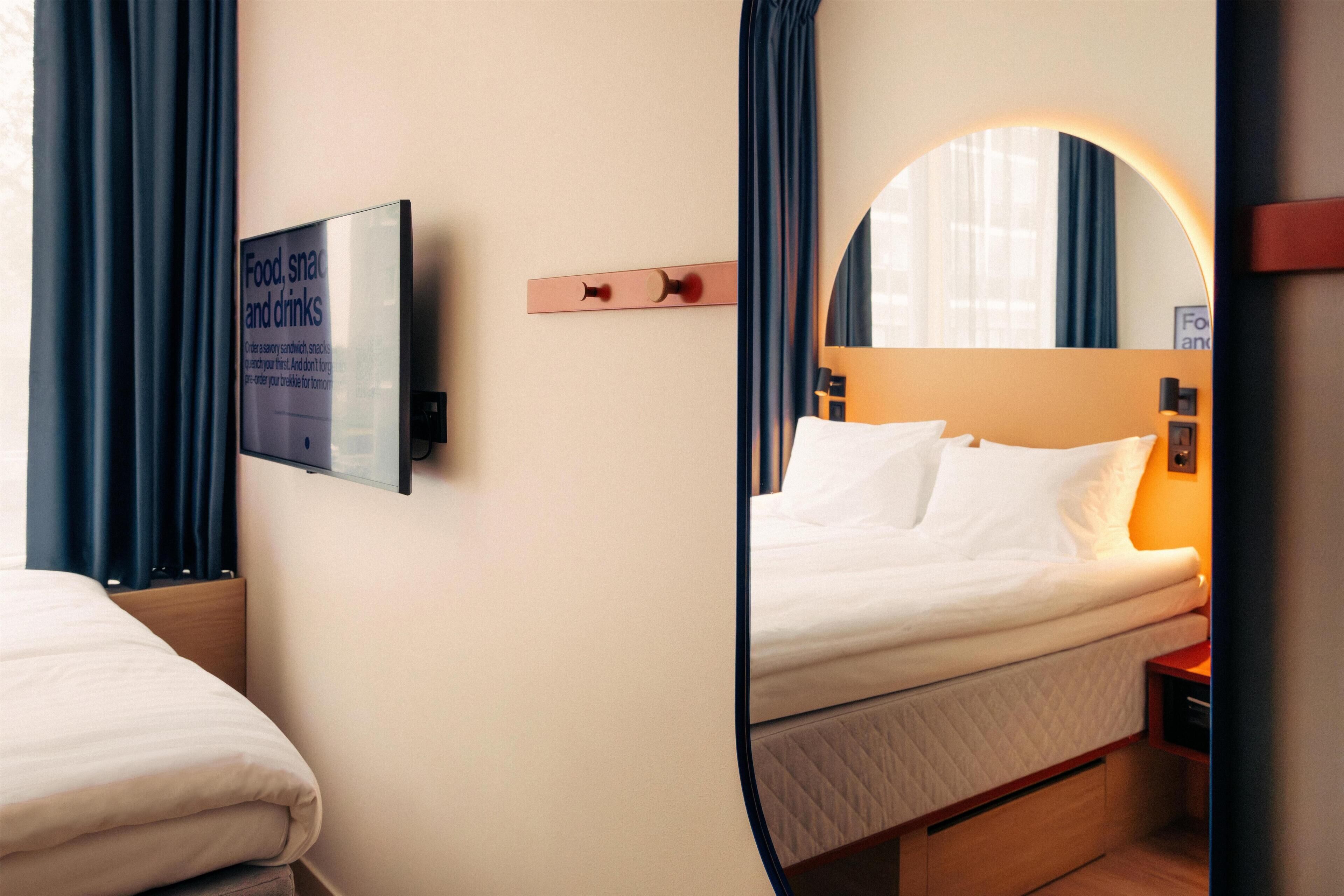 A hotel room with a bed, a bedside table, a mirror, and a sign about food, snacks, and drinks.