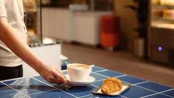 Eine Person serviert eine Tasse Kaffee und ein Croissant auf einem blau gefliesten Tisch.