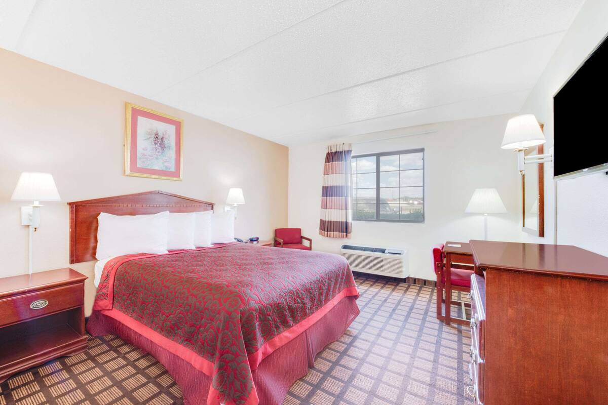 A hotel room with a large bed, two bedside tables, a desk, a chair, a window with curtains, and a wall-mounted TV.