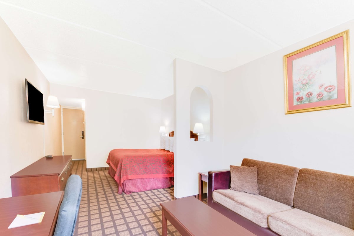 A hotel room with a bed, a sofa, a desk, a TV, and a painting on the wall.