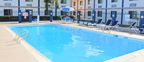 A swimming pool with lounge chairs and umbrellas, surrounded by a fence and buildings with a blue roof.