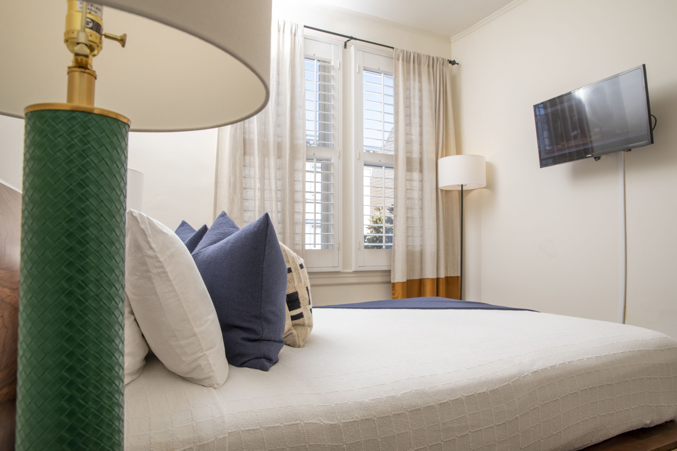 A hotel room with a bed, a green textured lamp, a television on the wall, and a window with curtains.