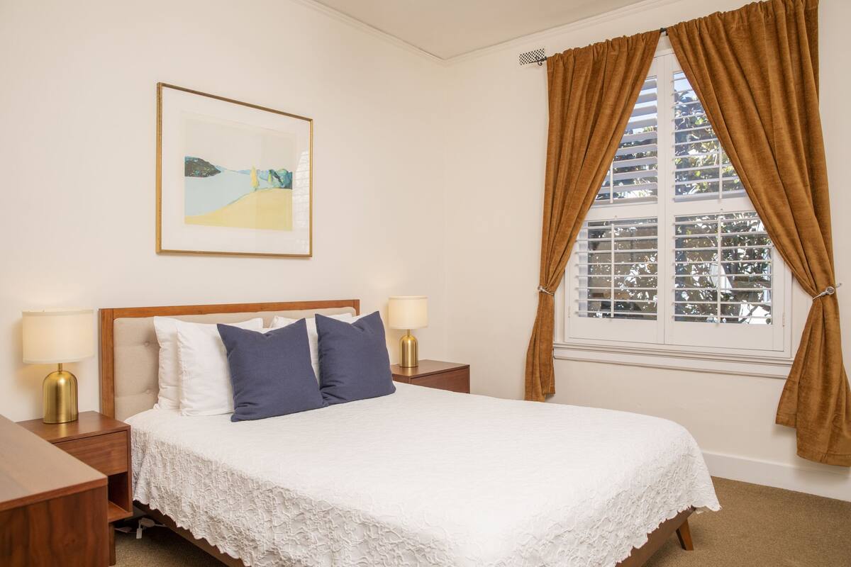 Quarto de hotel bem arrumado com uma cama, duas mesas de cabeceira, um abajur, um quadro na parede e uma janela com cortinas.