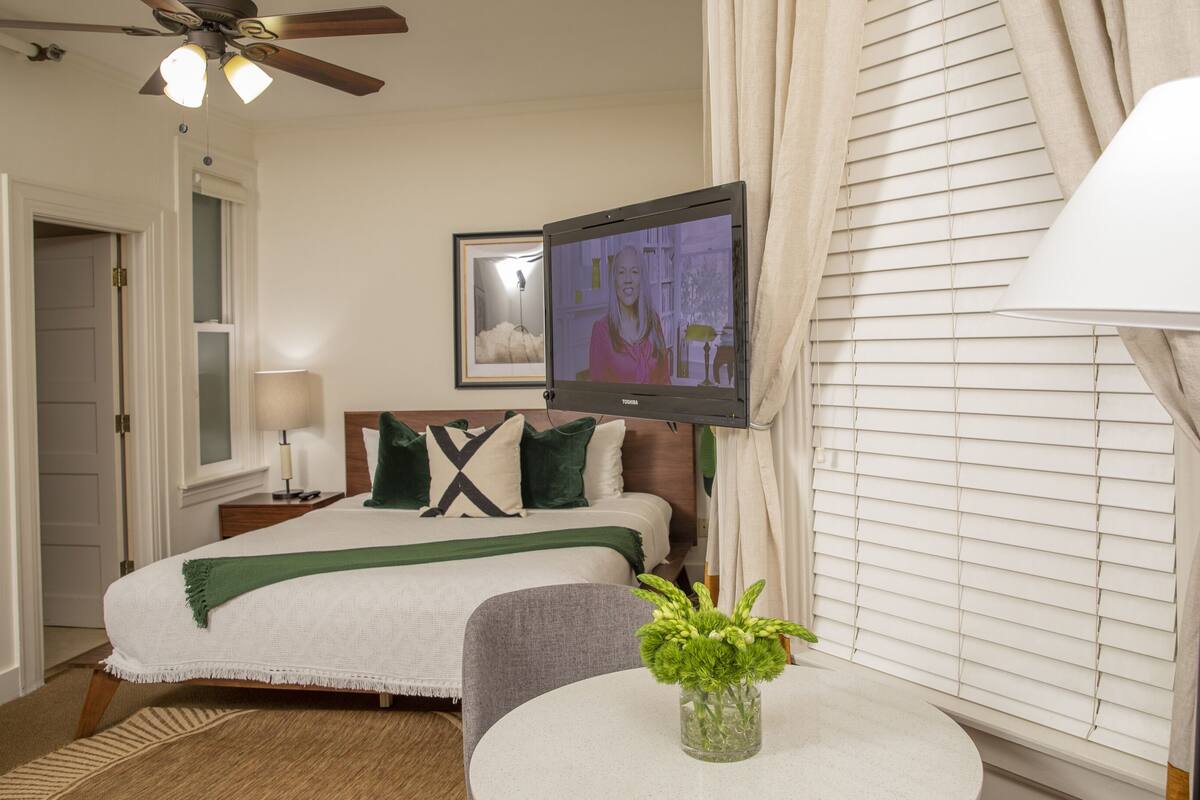 Quarto de hotel com cama, televisão na parede, mesa de cabeceira com abajur, janela com persianas e uma pequena mesa redonda com um vaso de flores.