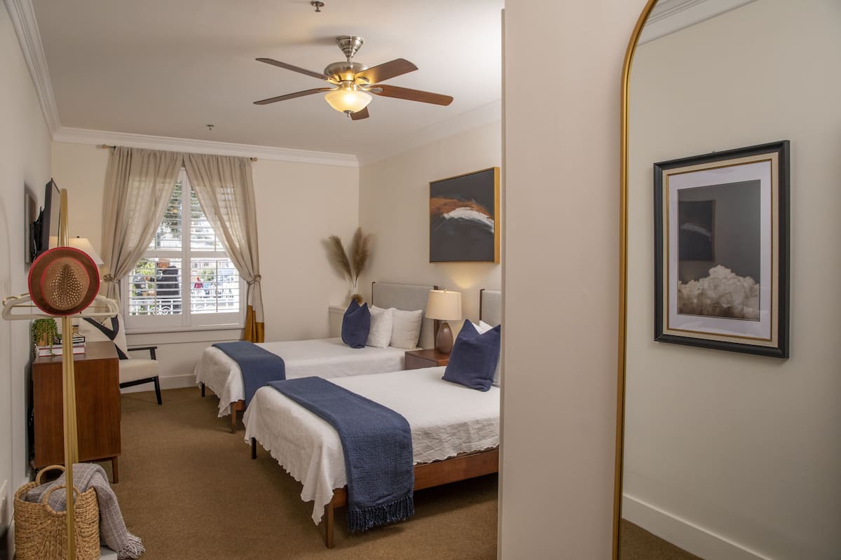Quarto de hotel com duas camas, um ventilador de teto, uma janela com cortinas, uma luminária, um quadro emoldurado e uma pequena mesa com uma planta.