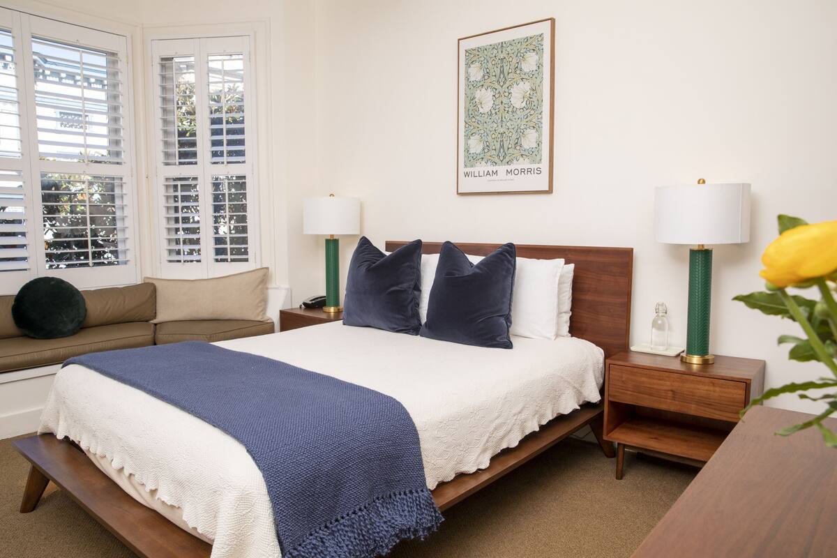 Quarto bem arrumado com cama, mesas de cabeceira, um banco e uma janela com persianas.