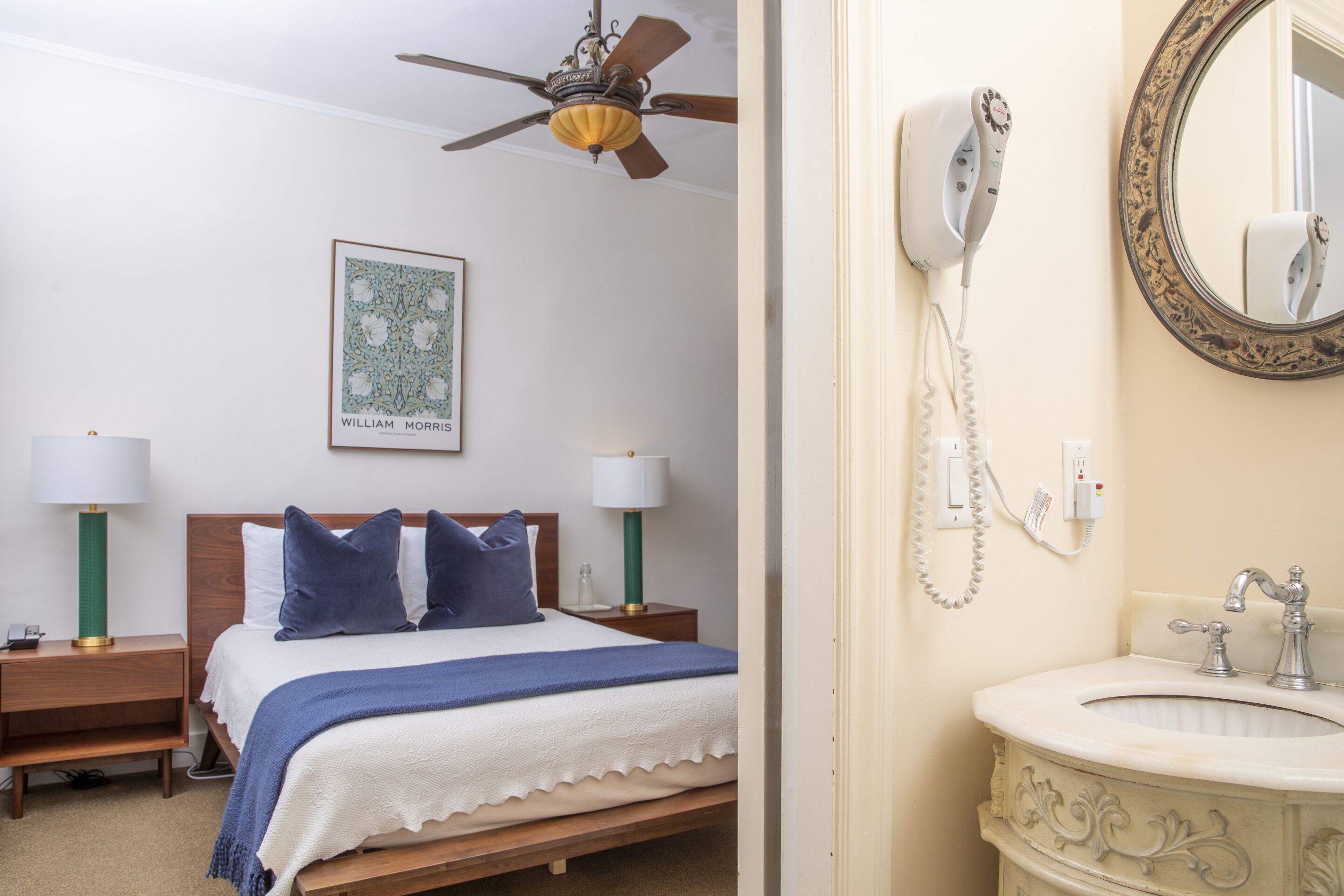 A bedroom with a bed, two bedside tables with lamps, a ceiling fan, a framed picture, and a bathroom with a sink, mirror, and hairdryer.