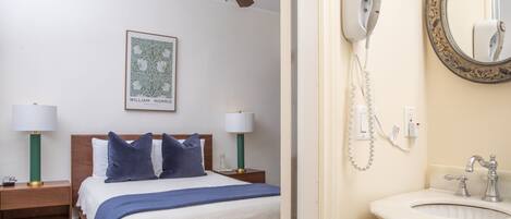 A bedroom with a bed, two bedside tables with lamps, a ceiling fan, a framed picture, and a bathroom with a sink, mirror, and hairdryer.