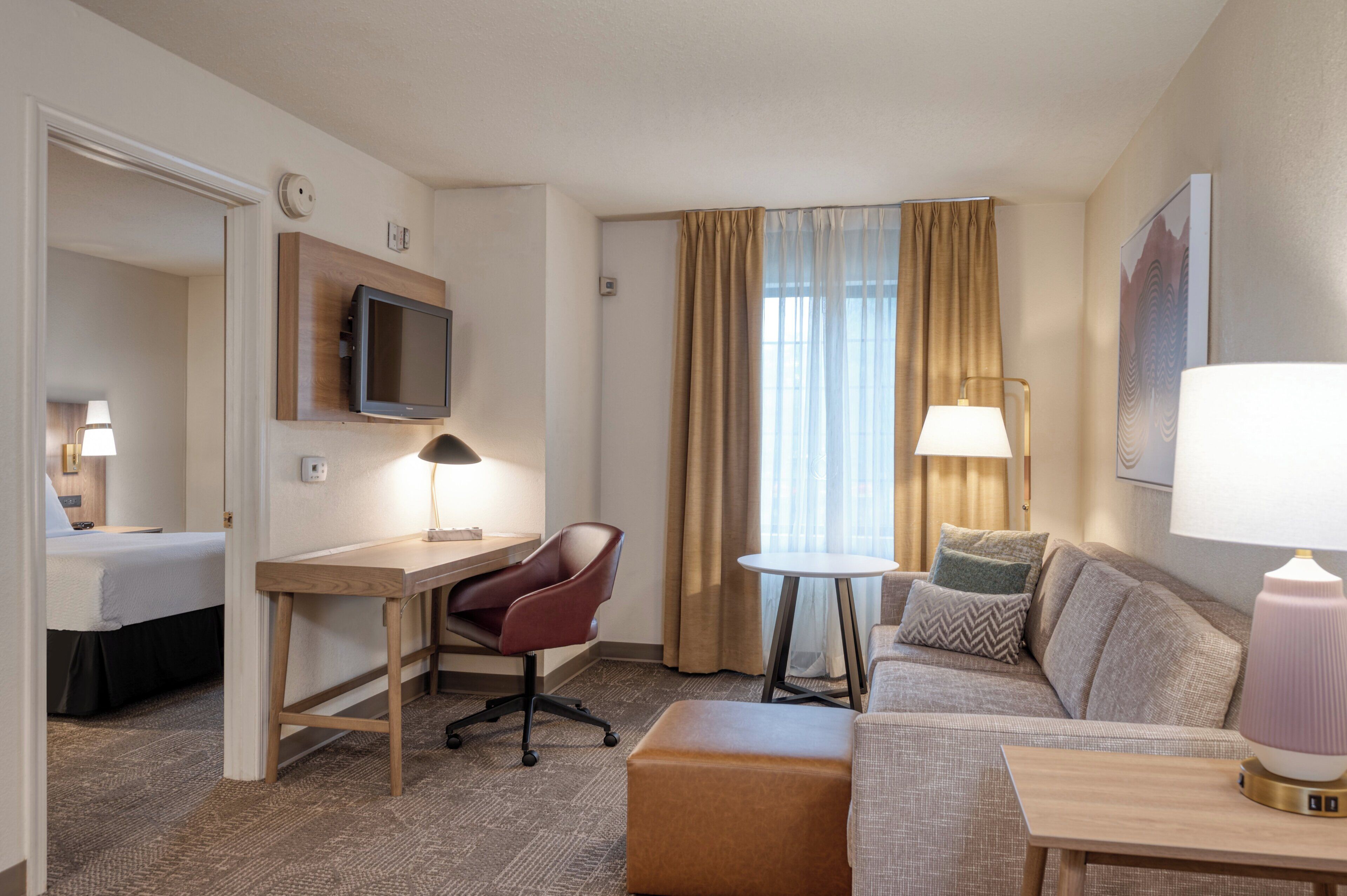 A hotel room with a desk, chair, sofa, ottoman, and two lamps.
