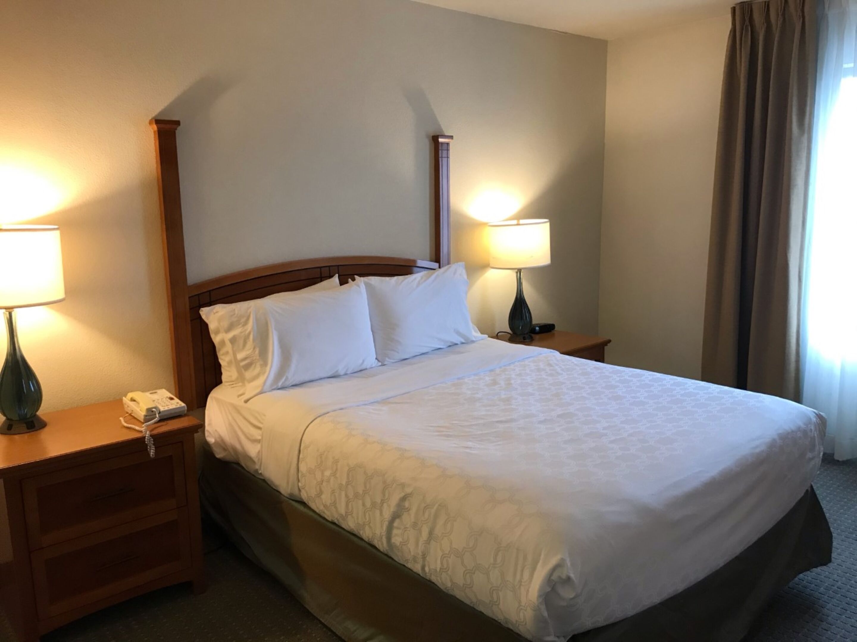 A neatly made bed with white linens, two bedside lamps, a wooden nightstand, and a telephone.