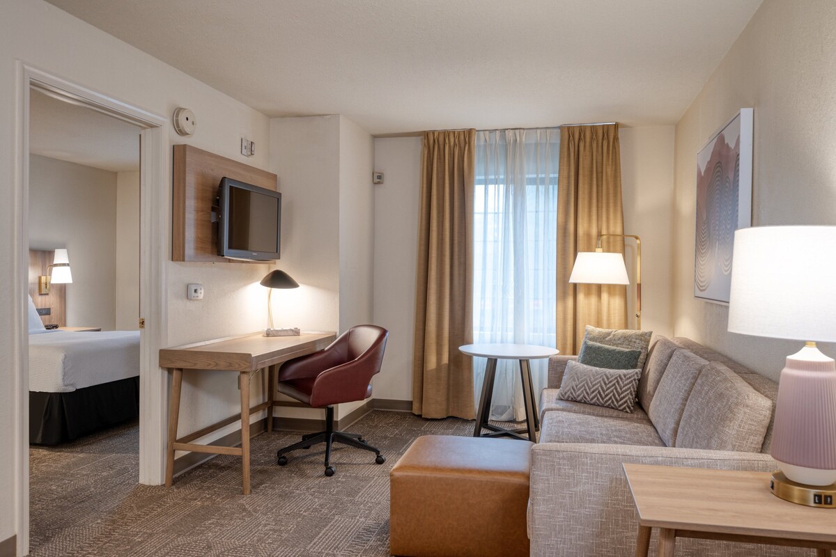 Ein Hotelzimmer mit einem Bett, einem Schreibtisch mit Stuhl, einem Sofa, einem kleinen Tisch und einem an der Wand befestigten Fernseher.
