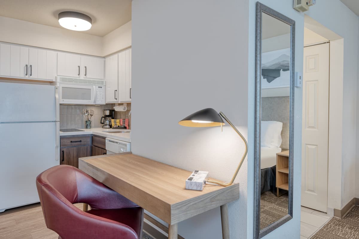 Une cuisine compacte avec des meubles blancs, un four à micro-ondes et une machine à café. On y trouve un bureau en bois avec une lampe, une chaise rouge et une petite boîte blanche.