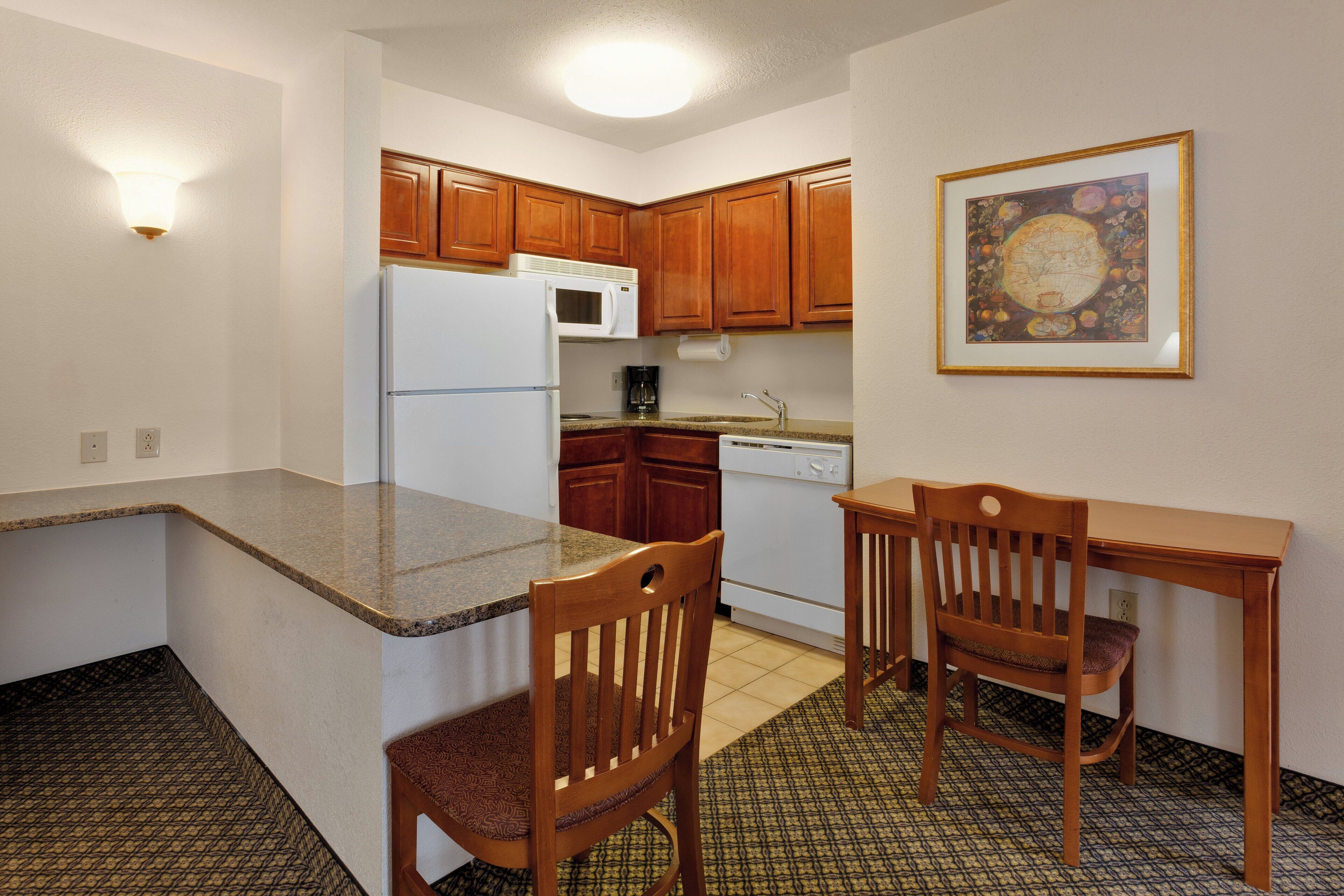 Cocina compacta con armarios de madera, un refrigerador blanco, microondas, cafetera y una mesa de comedor con dos sillas.