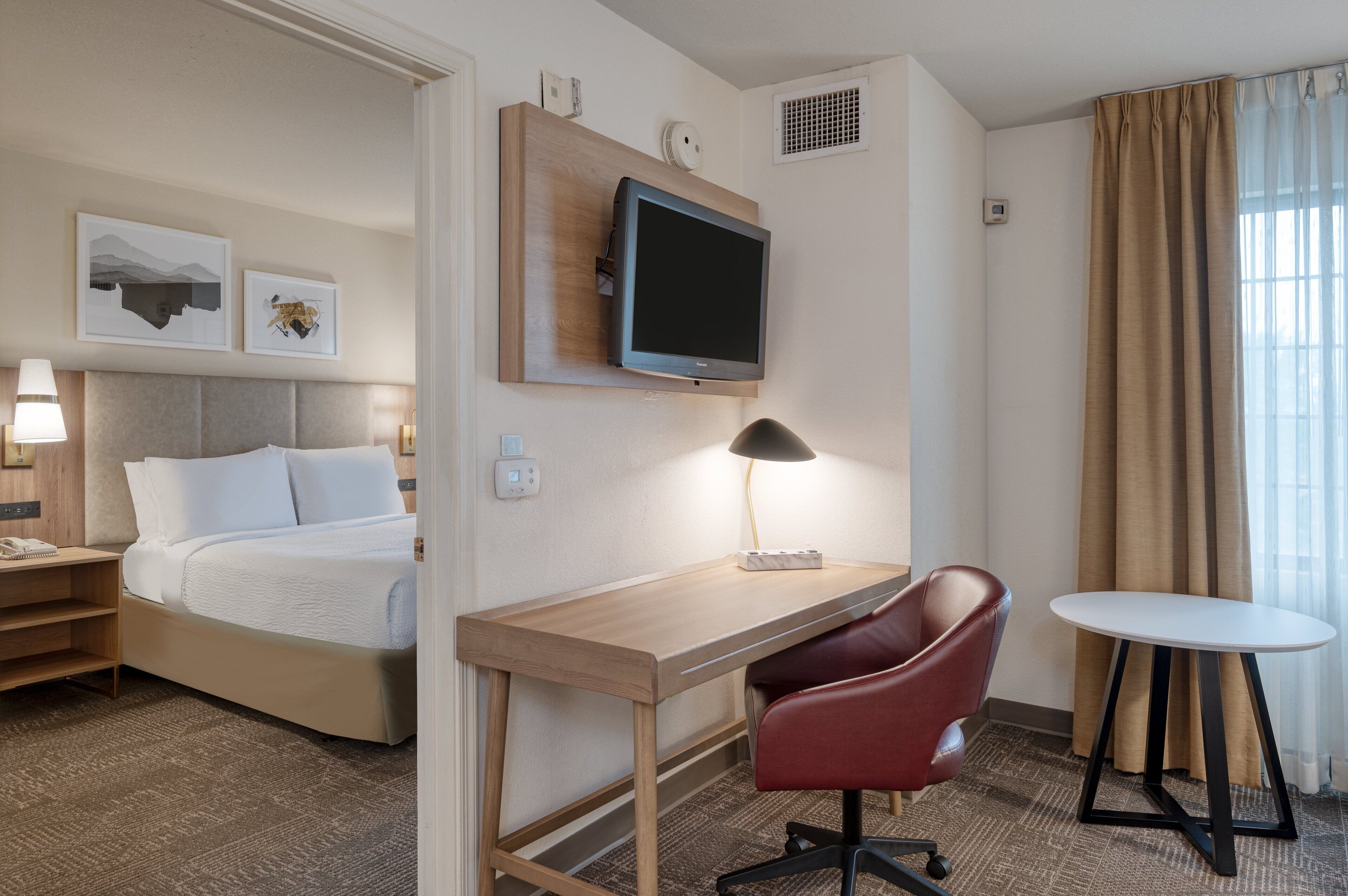 A hotel room with a bed, desk, chair, TV, and artwork on the wall.