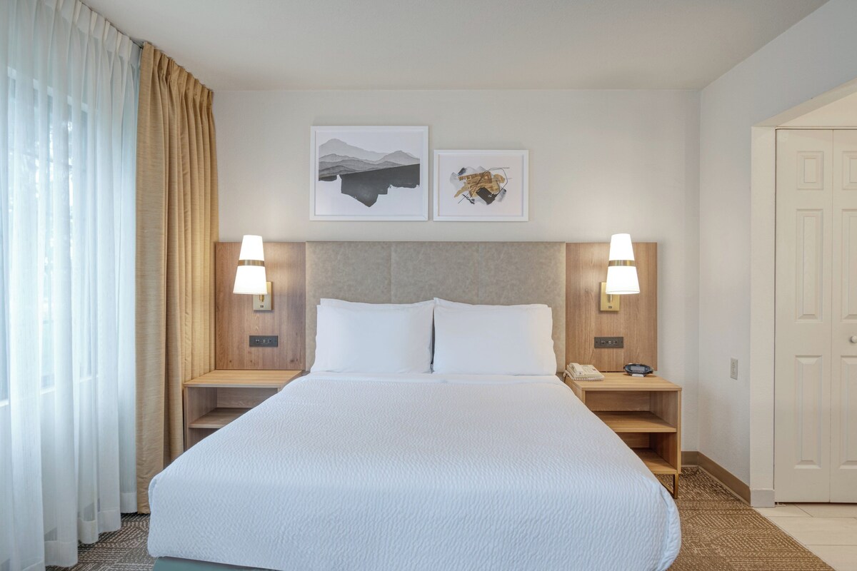 Une chambre d’hôtel avec un grand lit, deux tables de chevet avec des lampes, une tête de lit en bois et deux tableaux encadrés au mur.