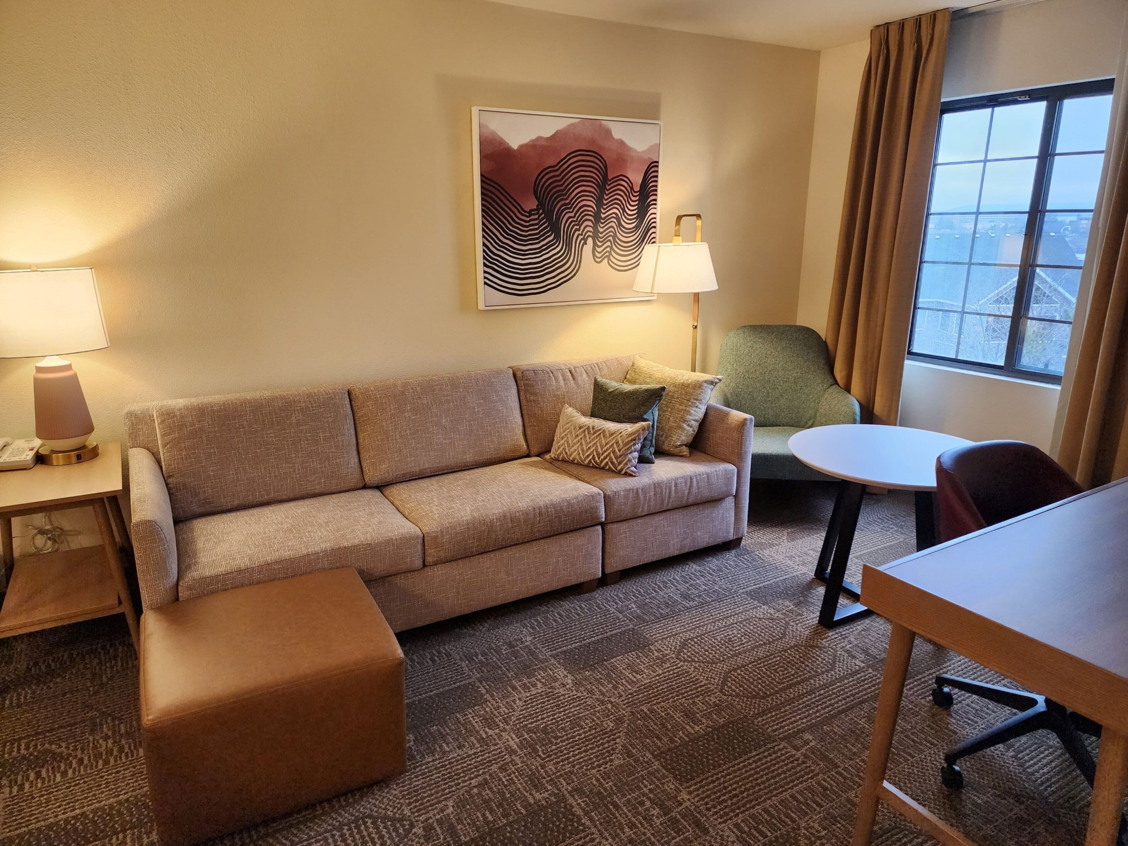 Hotellihuoneessa on sohva, rahi, pöytä ja tuoleja. Seinällä on taulu ja ikkunassa verhot.