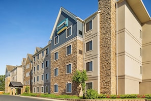 Un hôtel à plusieurs étages, doté d’une façade en pierre, d’une enseigne bien visible indiquant « Staybridge Suites », et d’un parterre aménagé devant l’entrée.