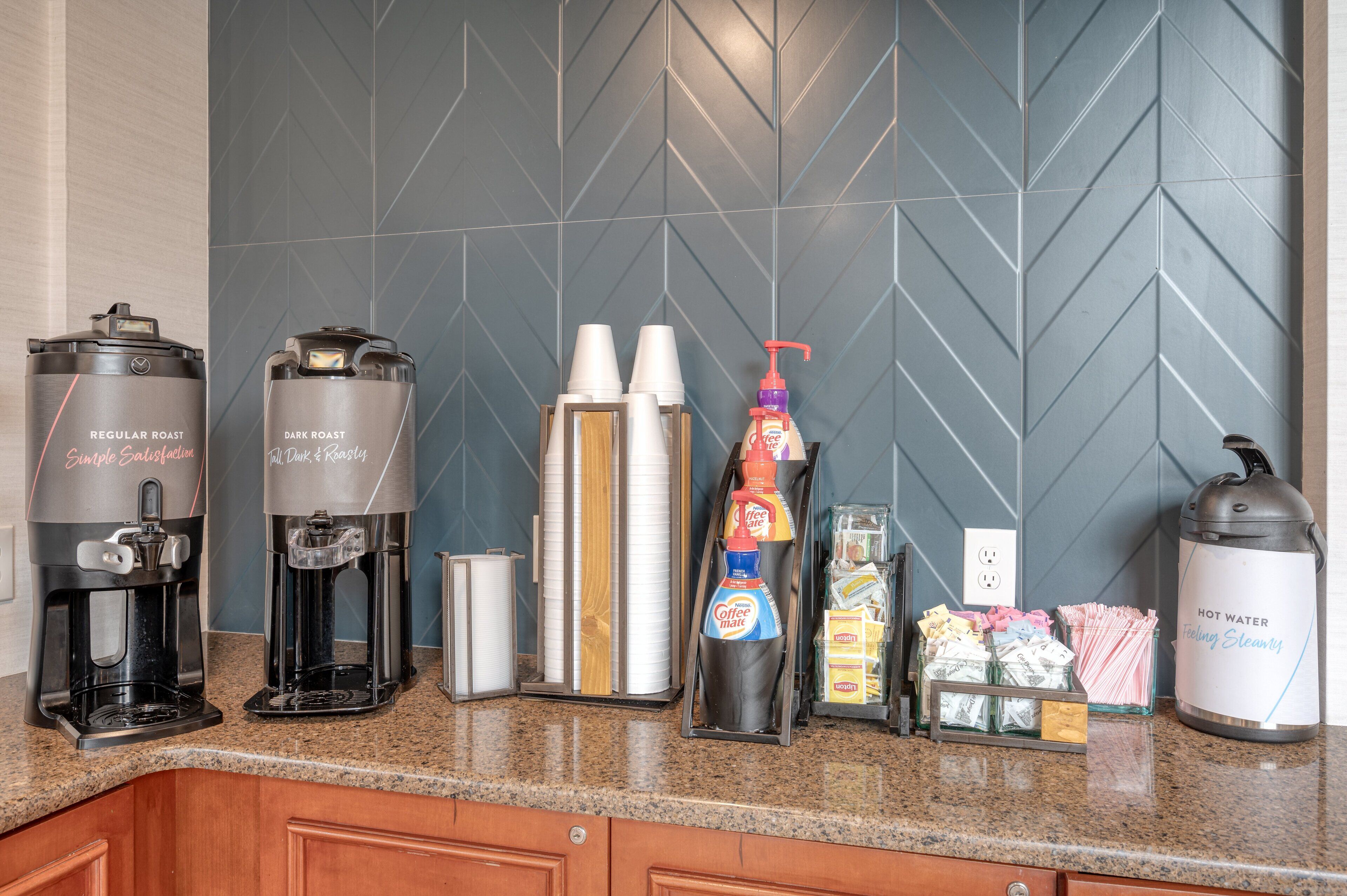 A coffee station with two coffee makers, disposable cups, a bottle of coffee creamer, and a hot water dispenser on a granite countertop.