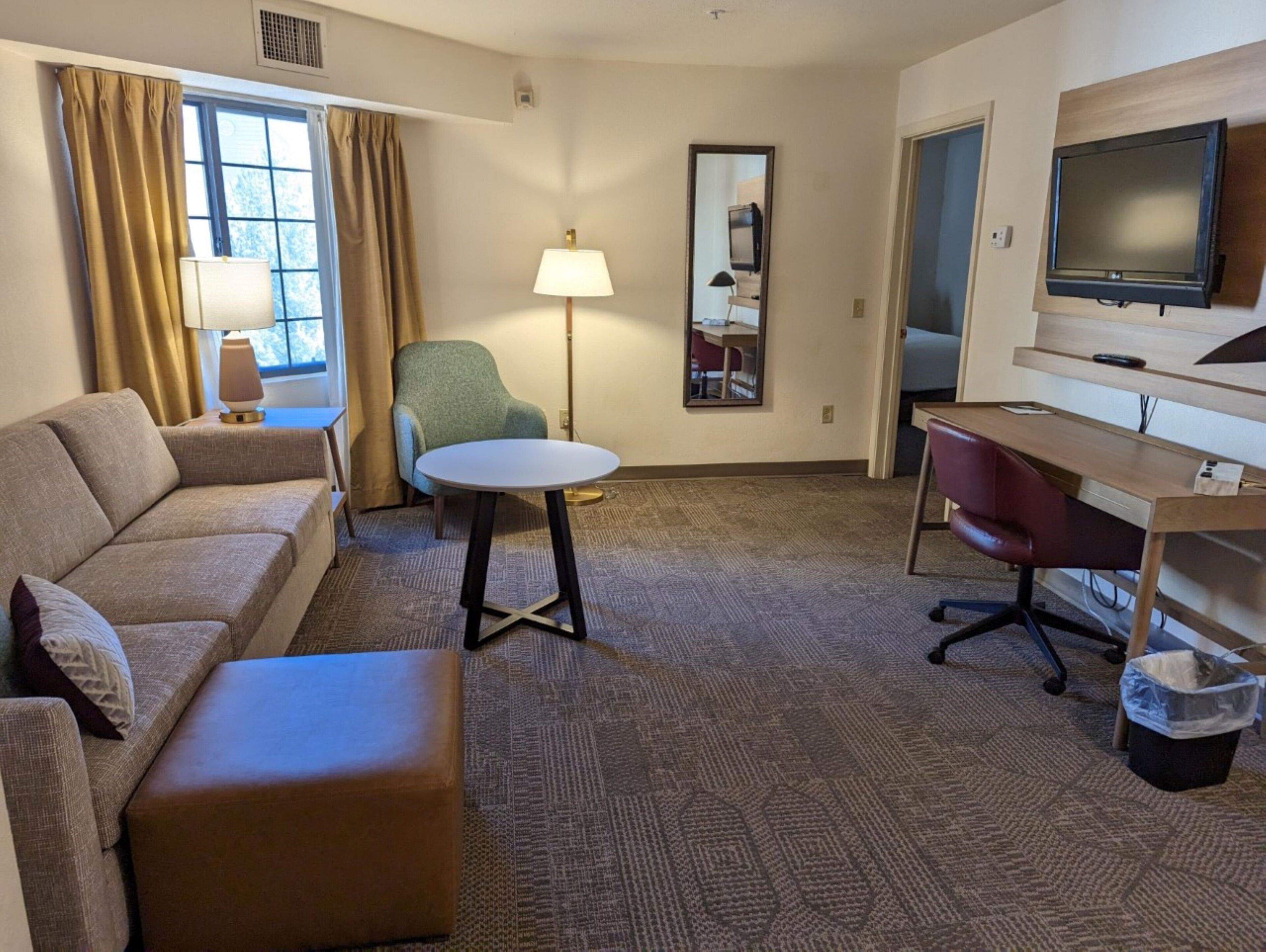A hotel room with a sofa, a small round table, a desk with a chair, a TV, and a lamp.