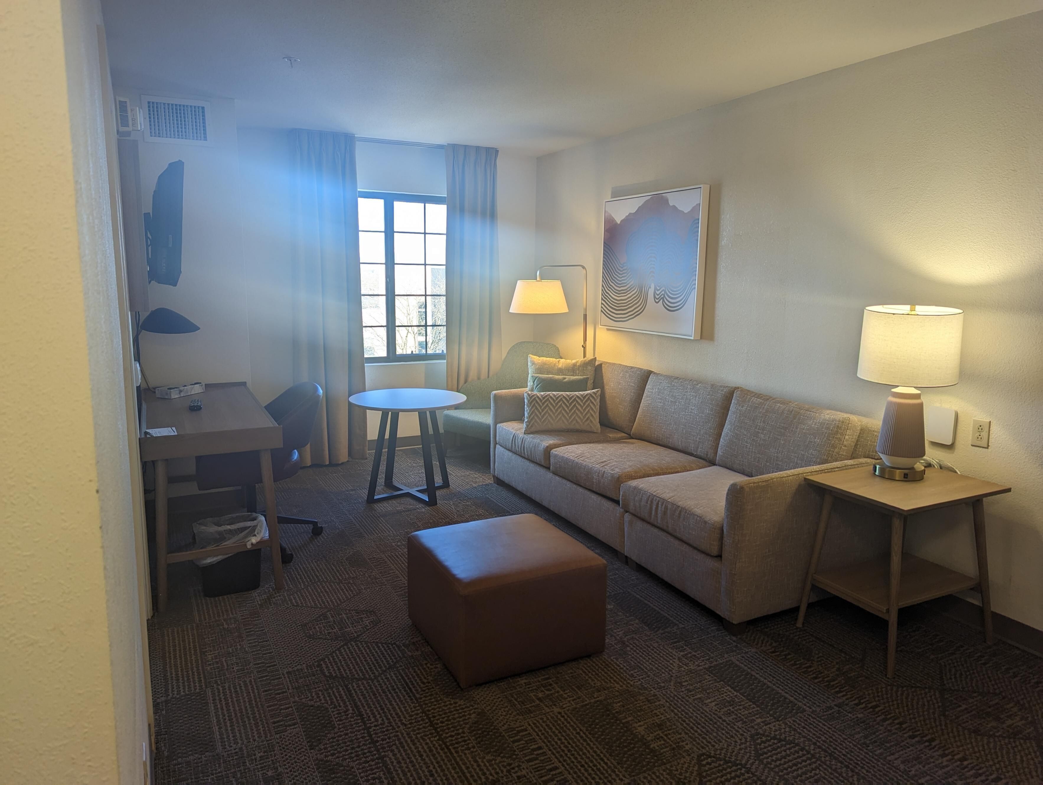 Habitación de hotel con sofá, una mesita, lámpara, silla, ventana con cortinas y suelo alfombrado.