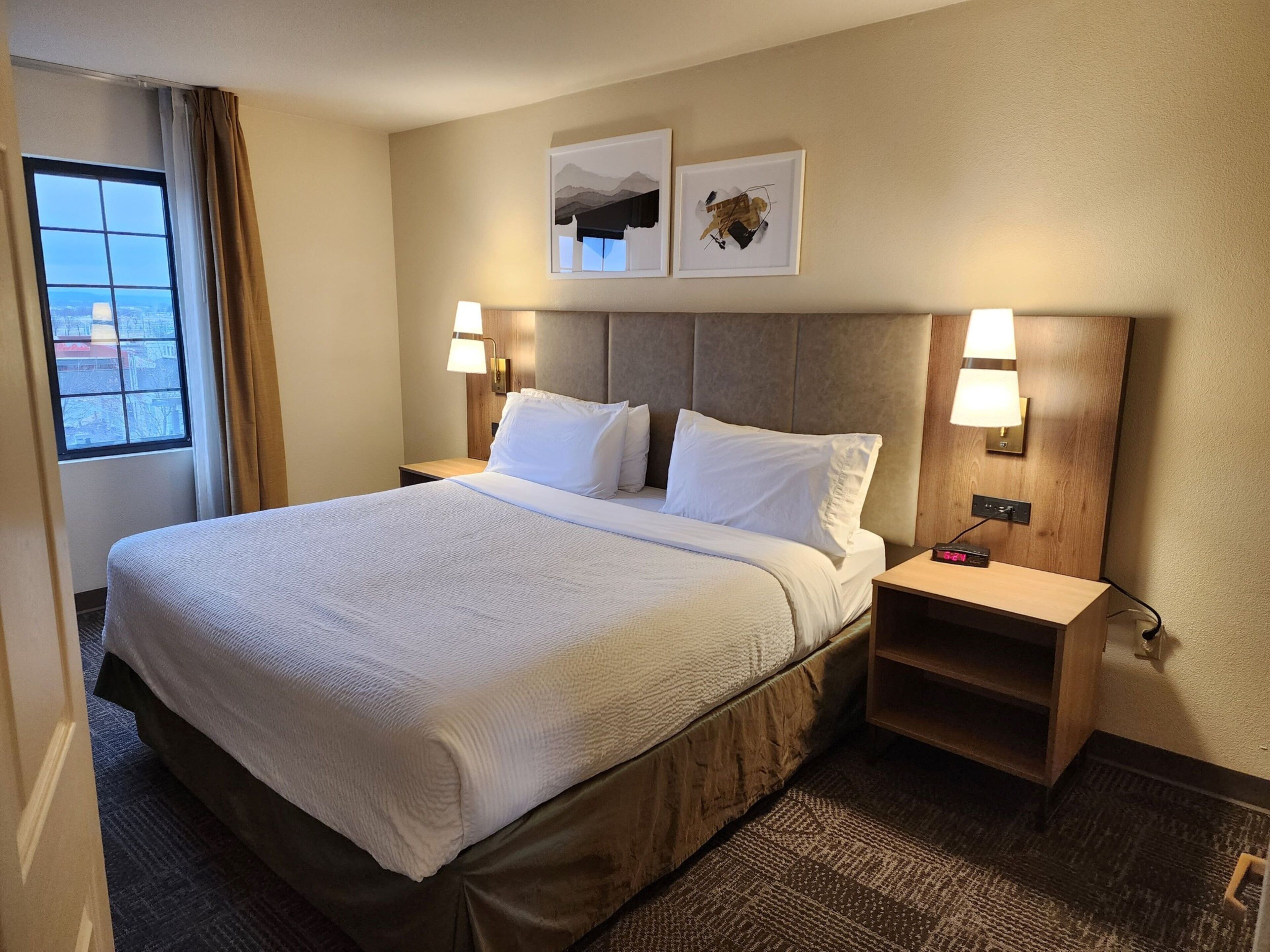 A hotel room with a large bed, two bedside tables, a nightstand, a window with curtains, and two wall-mounted lamps.