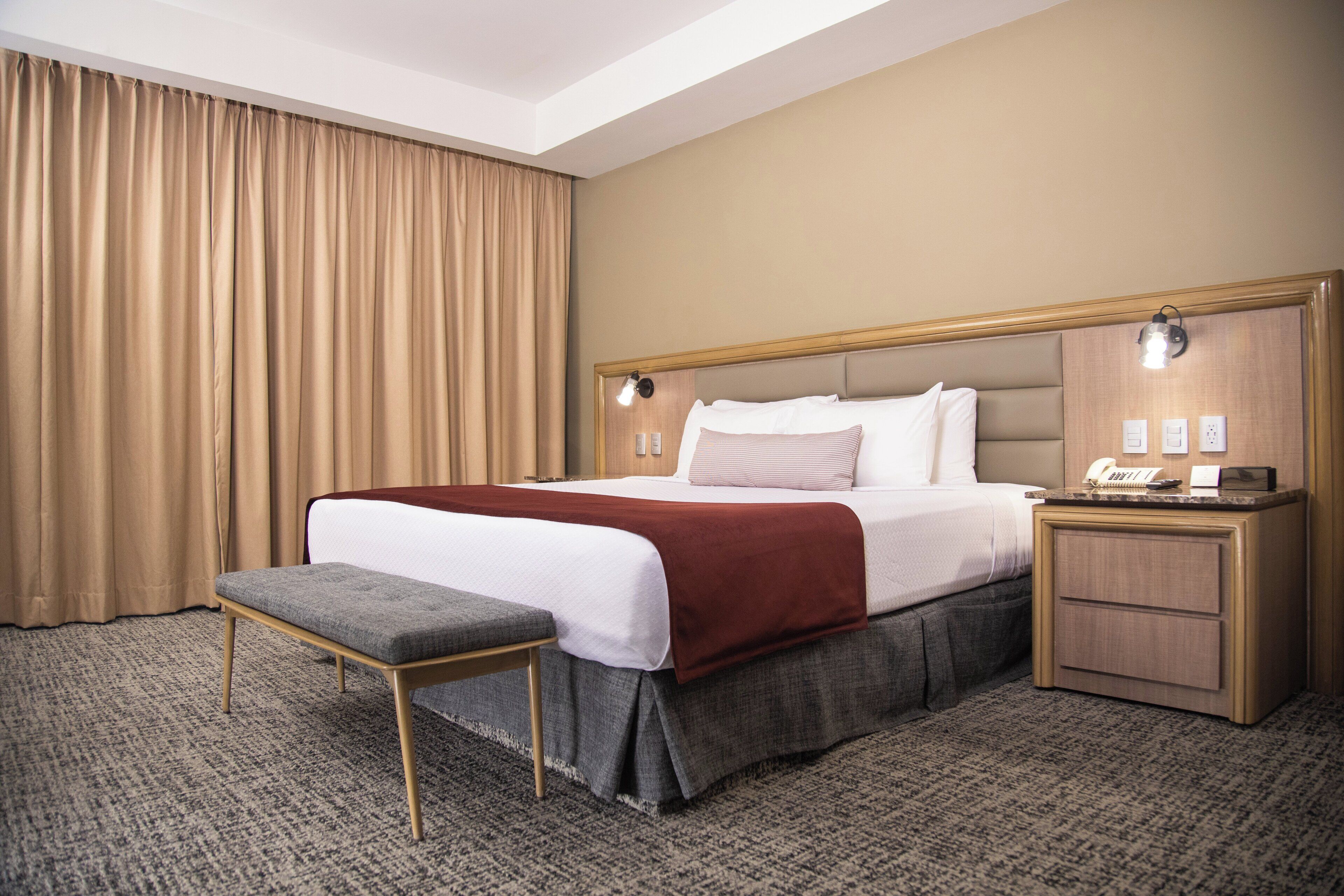 Una habitación de hotel con una cama grande, una mesita de noche, un banco y una ventana con cortinas.