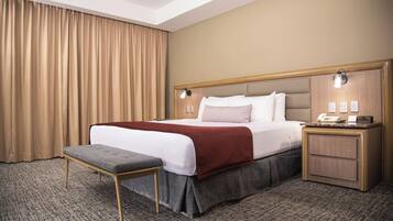 Una habitación de hotel con una cama grande, una mesita de noche, un banco y una ventana con cortinas.