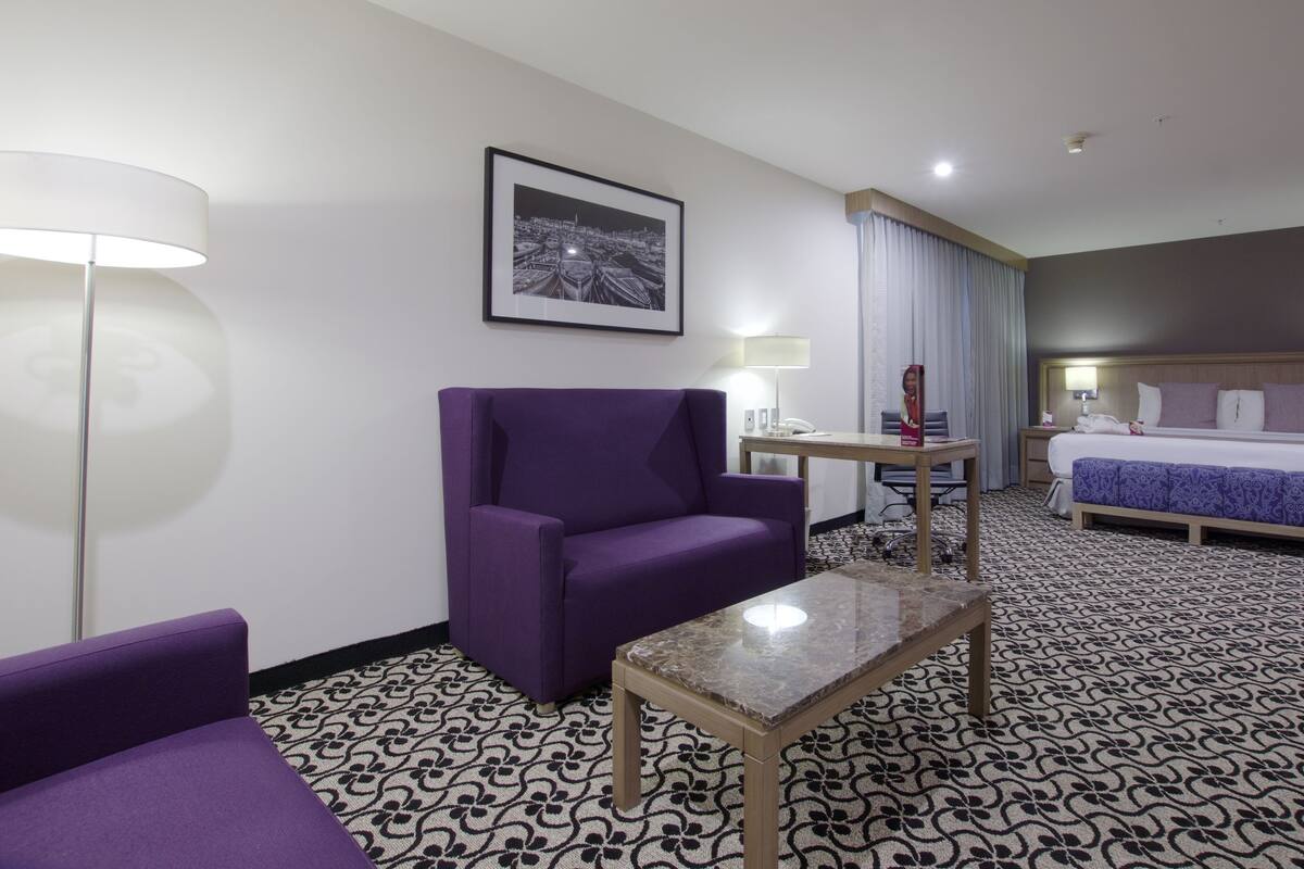 Una habitación de hotel con un sillón púrpura, una mesita, una cama con edredón azul, un cuadro en la pared y una alfombra estampada.