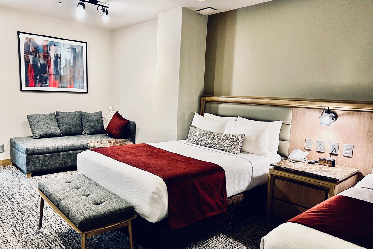 A hotel room with a bed, a sofa, a bench, a nightstand, a lamp, and a painting on the wall.