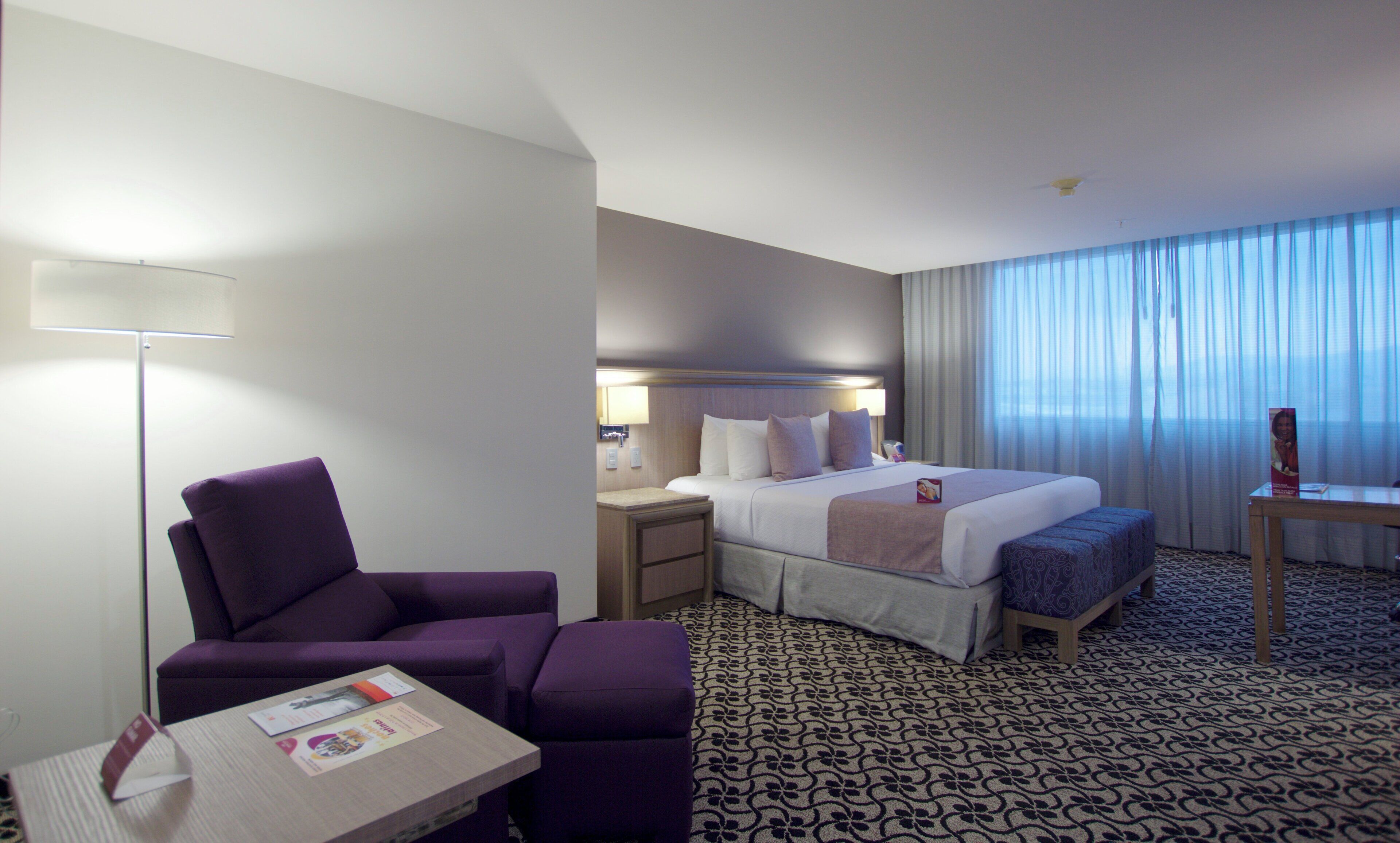 A hotel room with a bed, a chair, a bedside table, and a lamp.