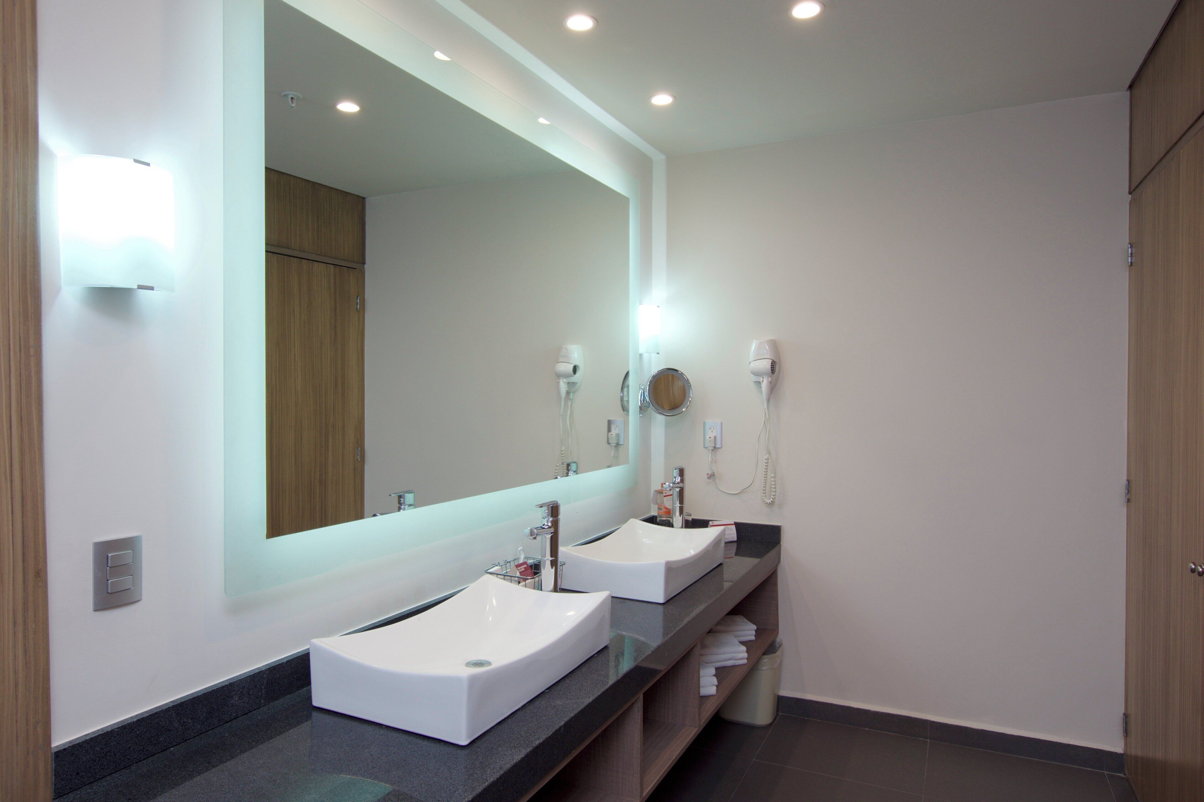 Une salle de bain moderne avec deux lavabos, un grand miroir et un sèche-cheveux fixé au mur.