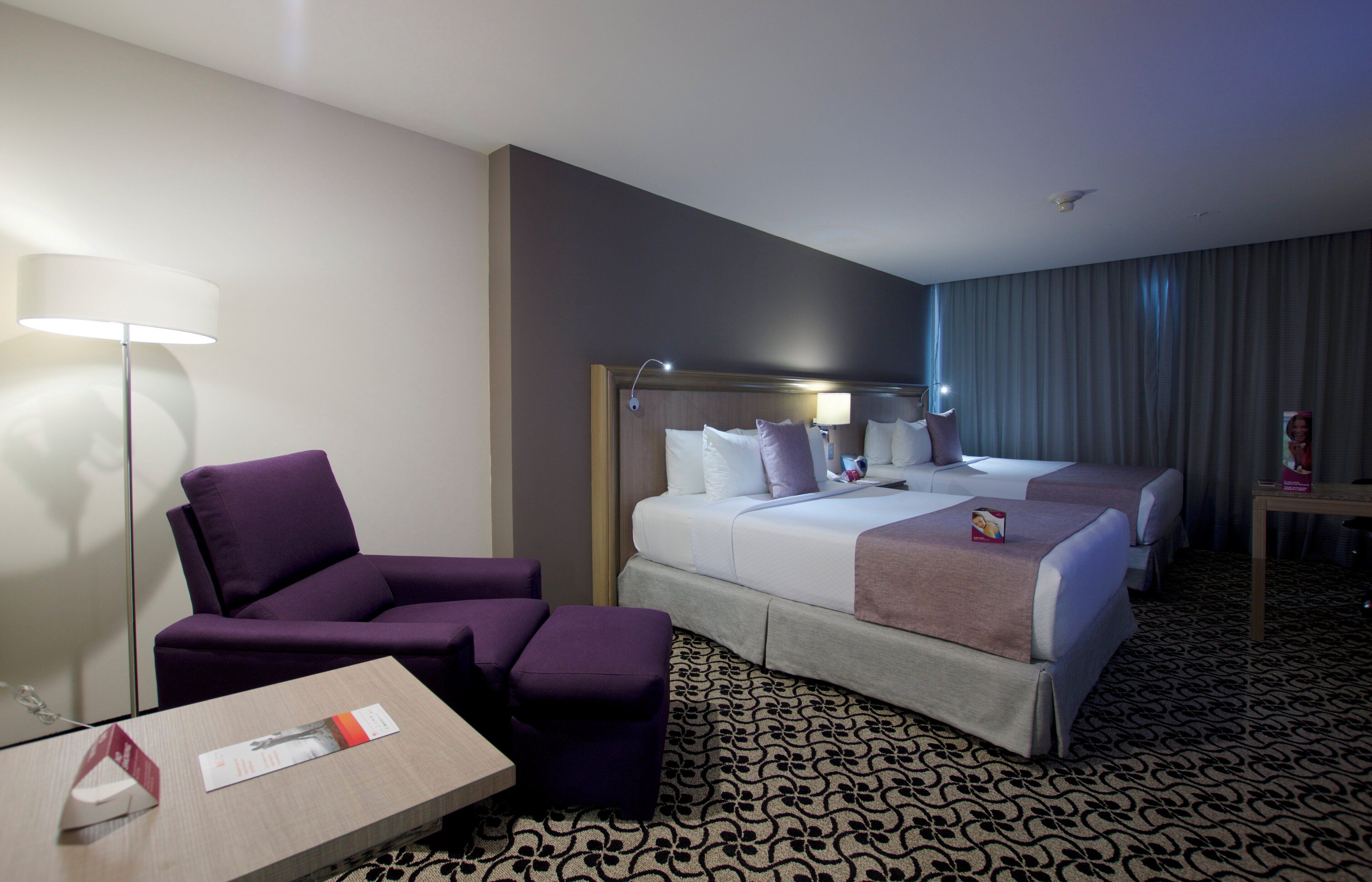 A hotel room with two beds, a purple armchair, a small table with a book and a remote control, and a patterned carpet.