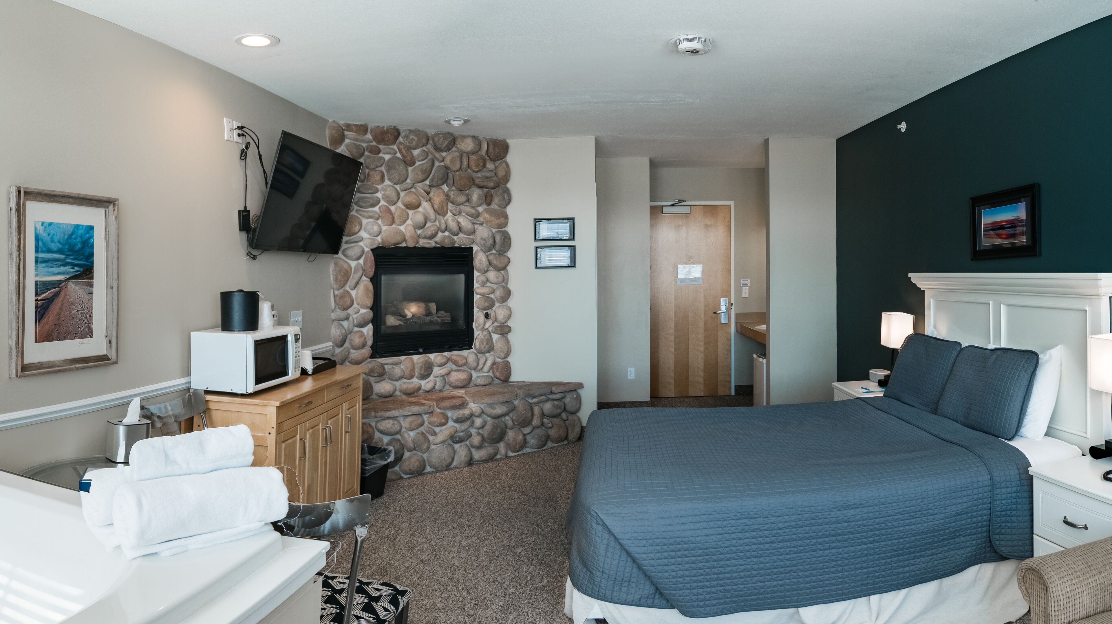 A hotel room with a stone fireplace, a bed with a blue quilt, a white bedside table, and a wooden nightstand.