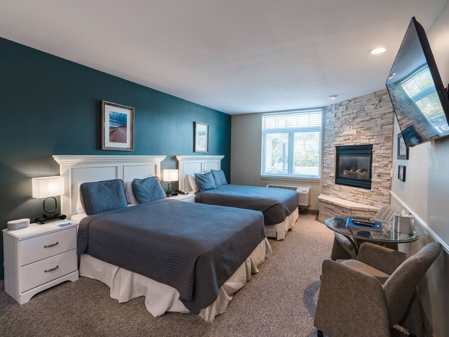 A hotel room with two beds, a stone fireplace, a TV mounted on the wall, and a window with blinds.