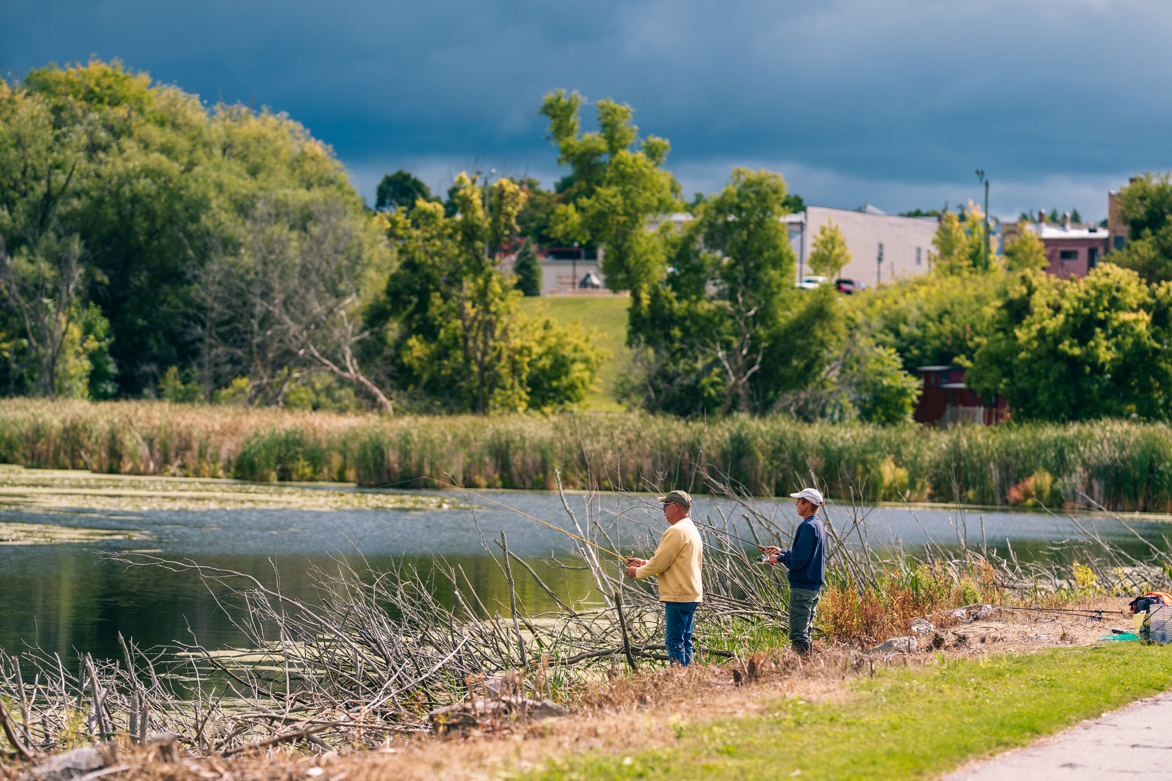 Two people fishing by a calm lake with a grassy bank and trees in the background.