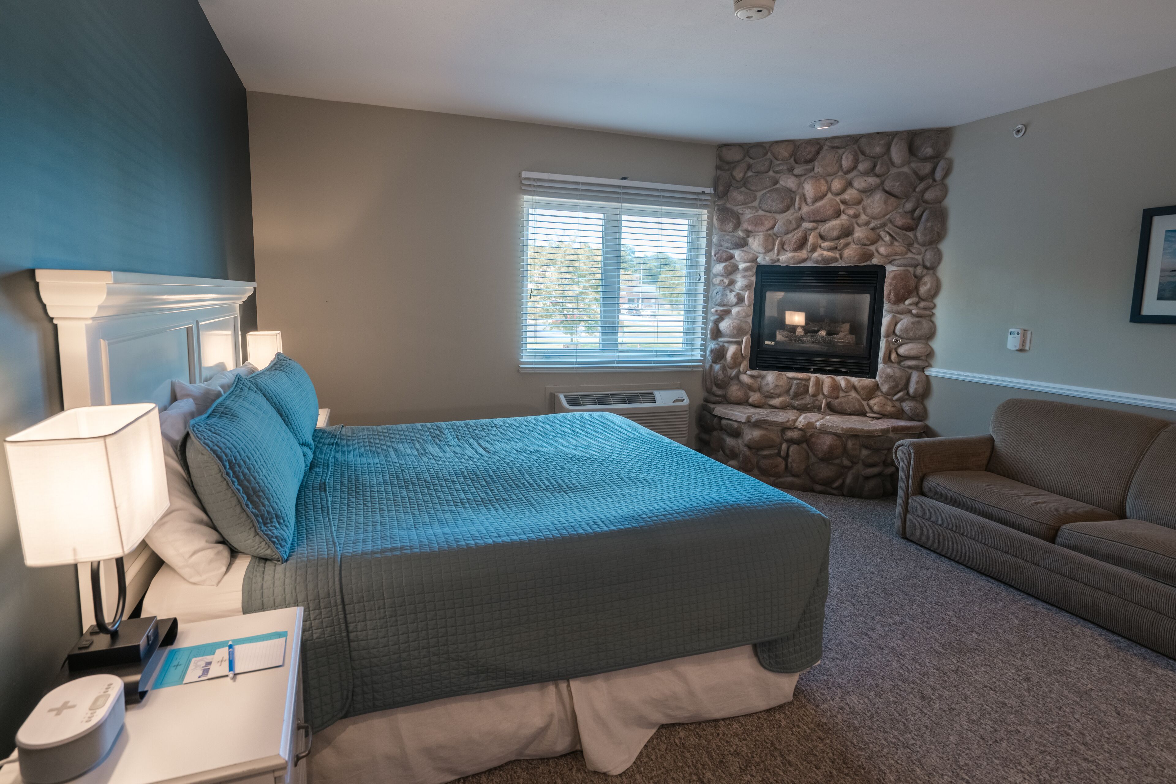 A bedroom with a bed, a nightstand, a lamp, a dresser, a stone fireplace, and a window with blinds.