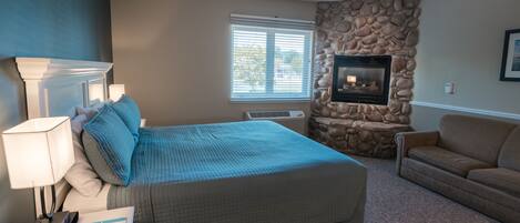 A bedroom with a bed, a nightstand, a lamp, a dresser, a stone fireplace, and a window with blinds.