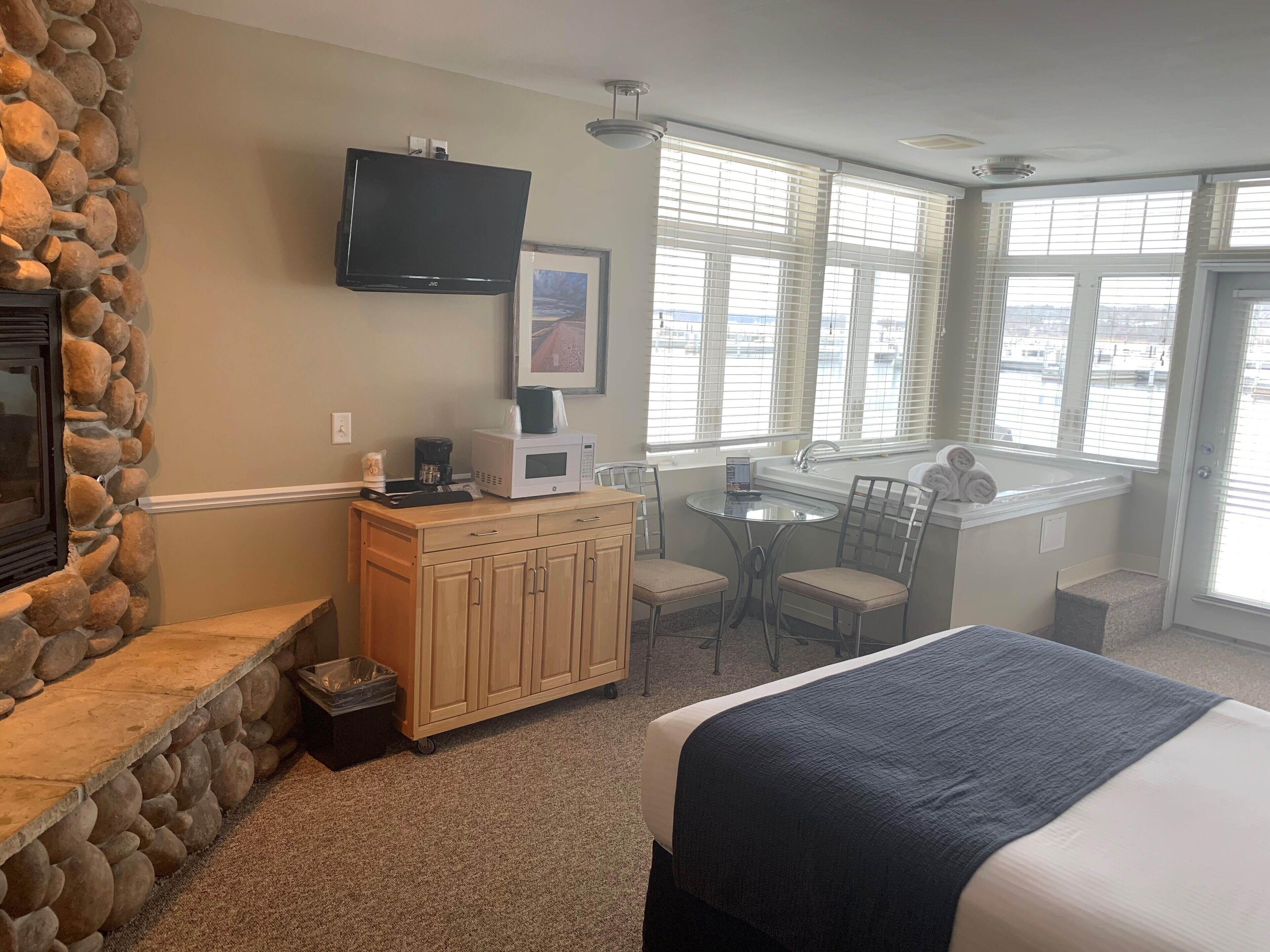 A hotel room with a stone wall, a flat-screen TV, a microwave, a coffee maker, a small table with chairs, a bed, and a window with blinds.